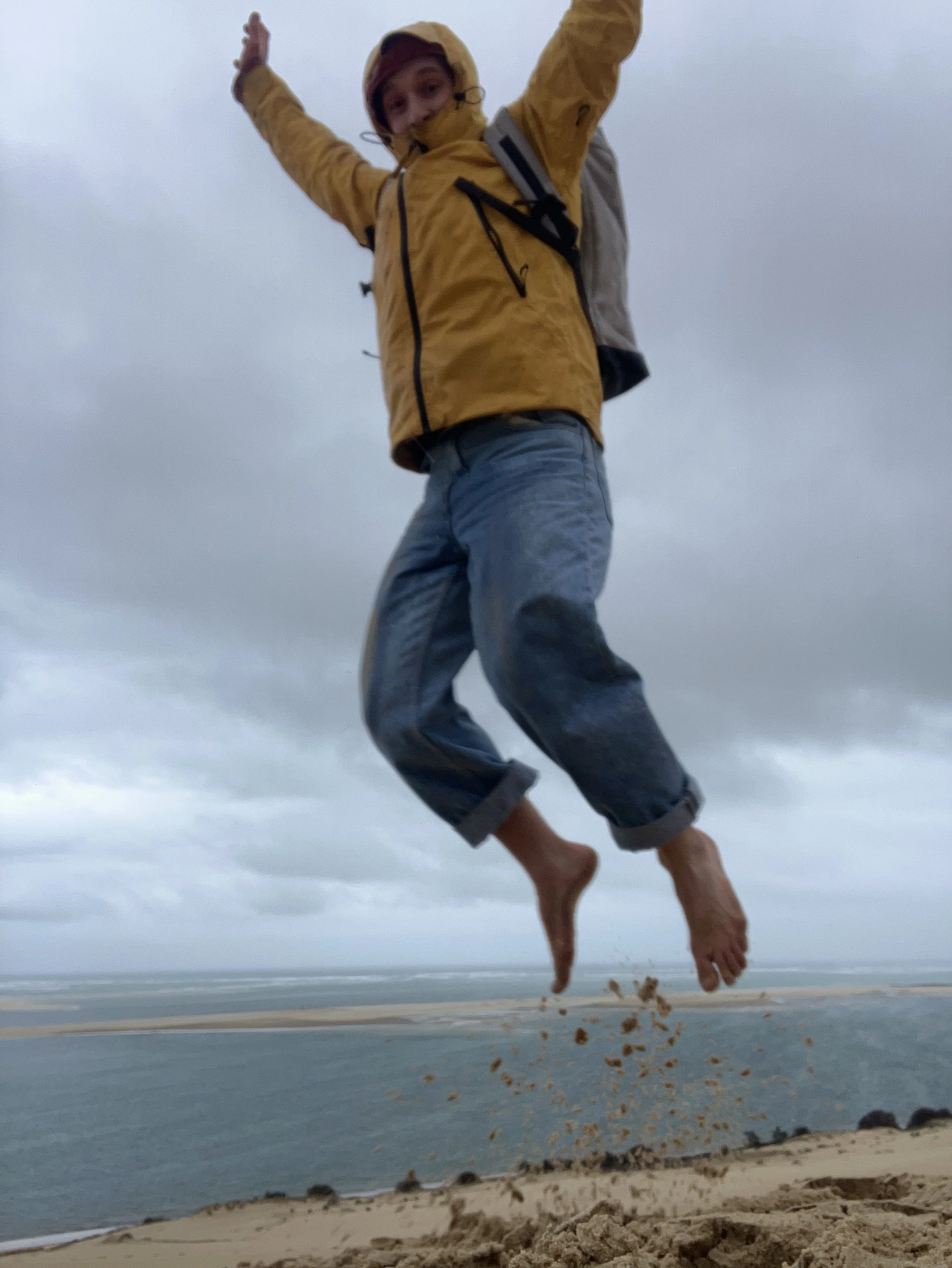 Une personne sautant au-dessus du sable avec la mer en arrière-plan, portant une veste jaune et un sac à dos, par temps nuageux.