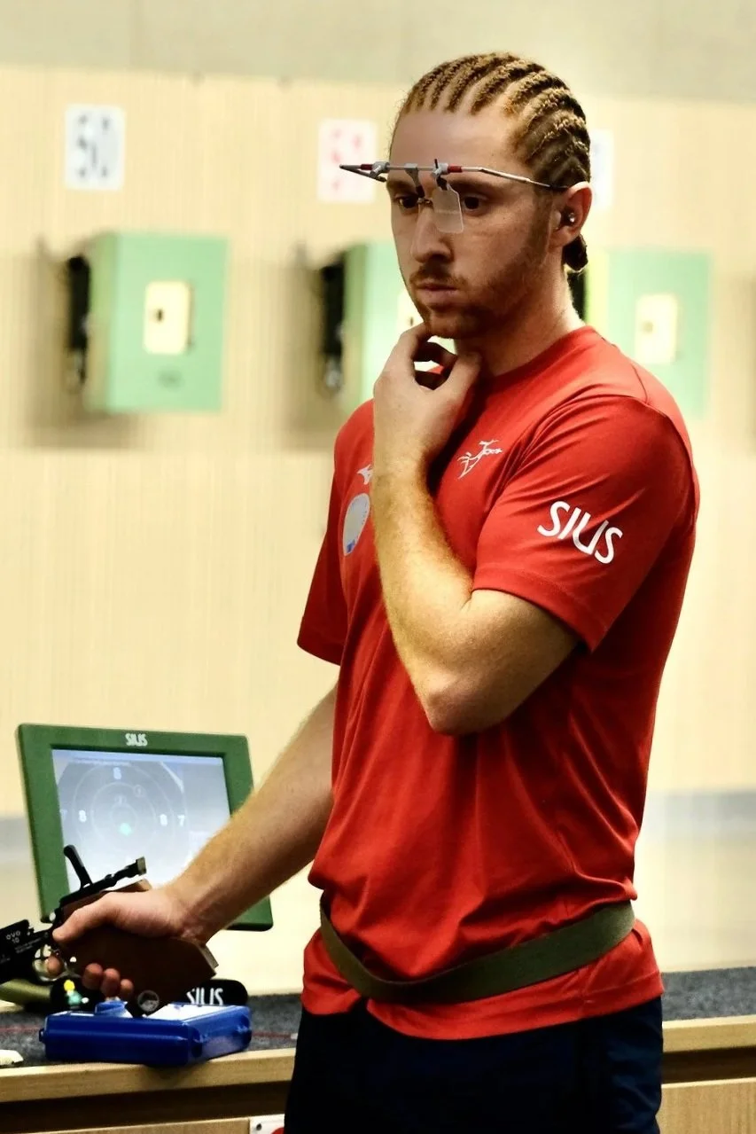 Un homme avec des cheveux en tresses portant un t-shirt rouge et des lunettes de visée électronique, semble concentré dans une salle d'entraînement de tir ou de tir sportif.