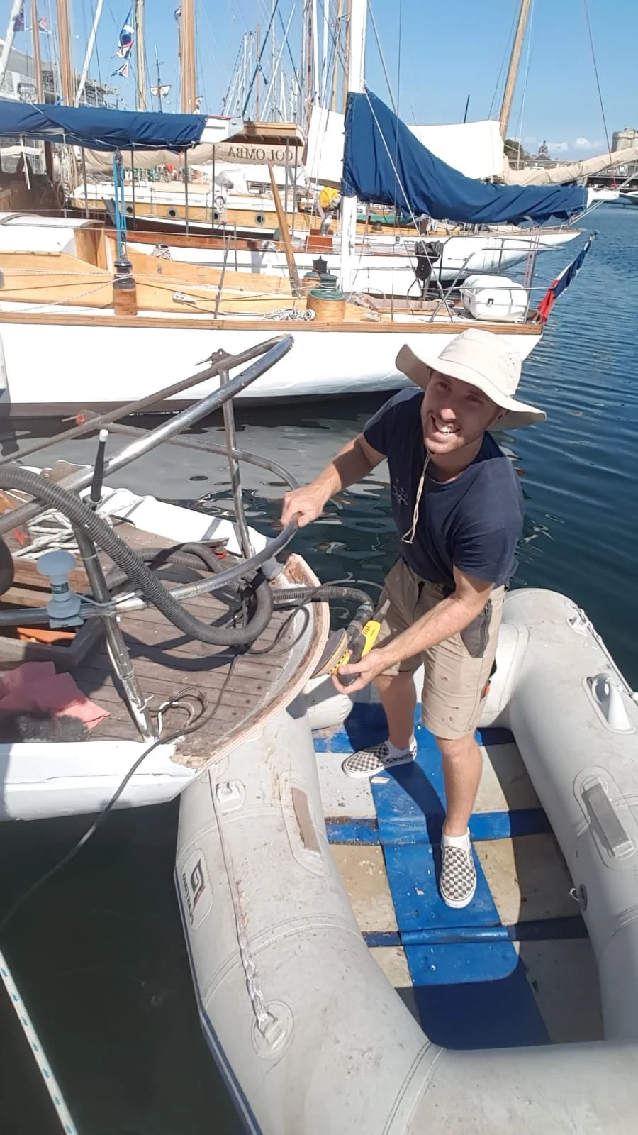 Un homme souriant, portant un chapeau beige, travaille sur un bateau à quai avec un tournevis dans la main, autour d'autres bateaux à voile au port.