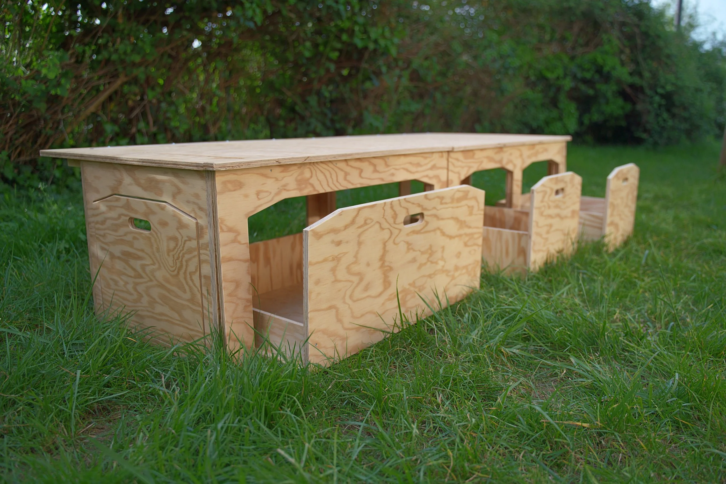 Une table en bois brut sur l'herbe, entourée de buissons verts.
