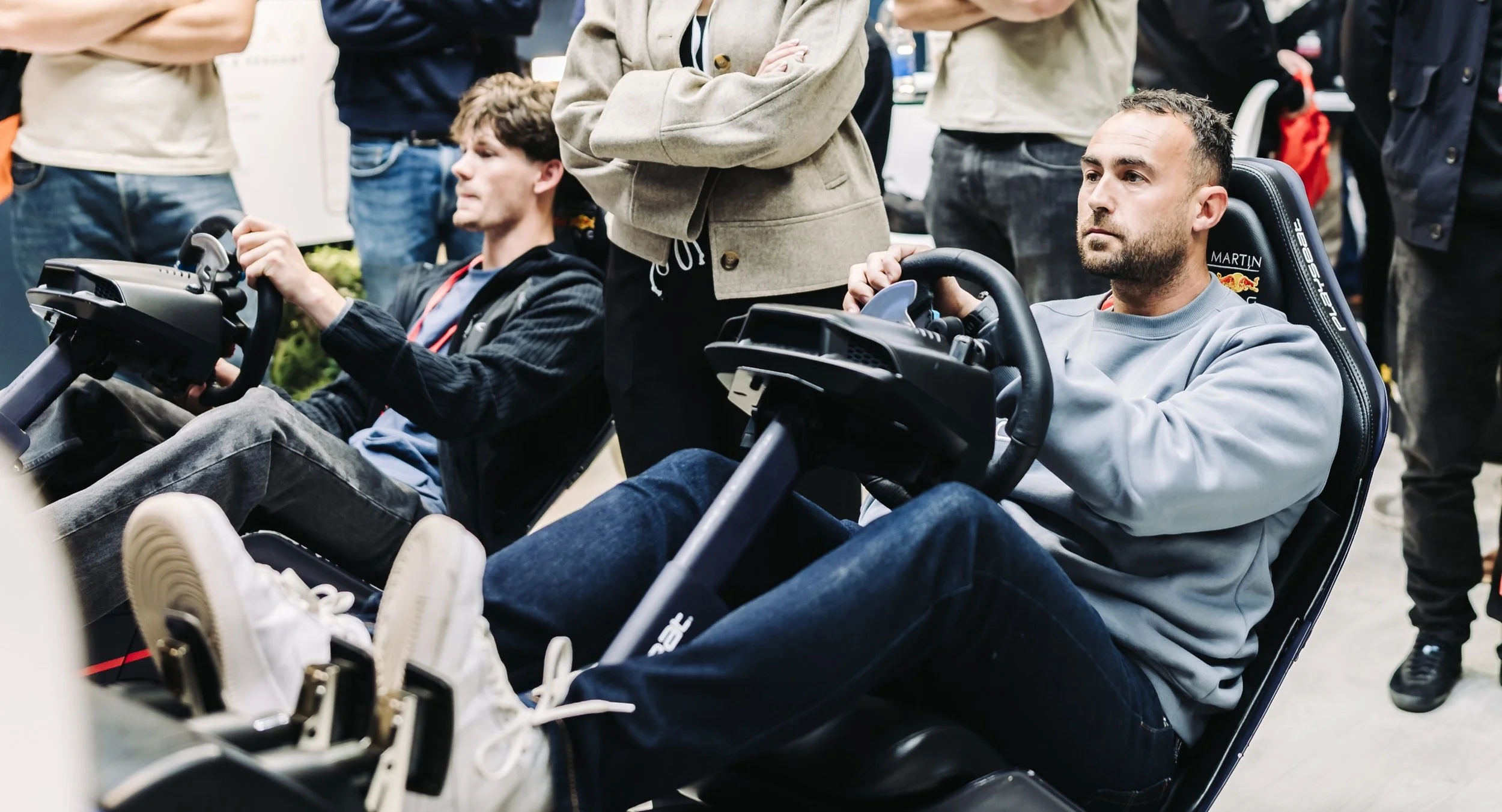 Two men seated in racing sim chairs, one steering with a serious expression, surrounded by standing people in a tech or gaming event.