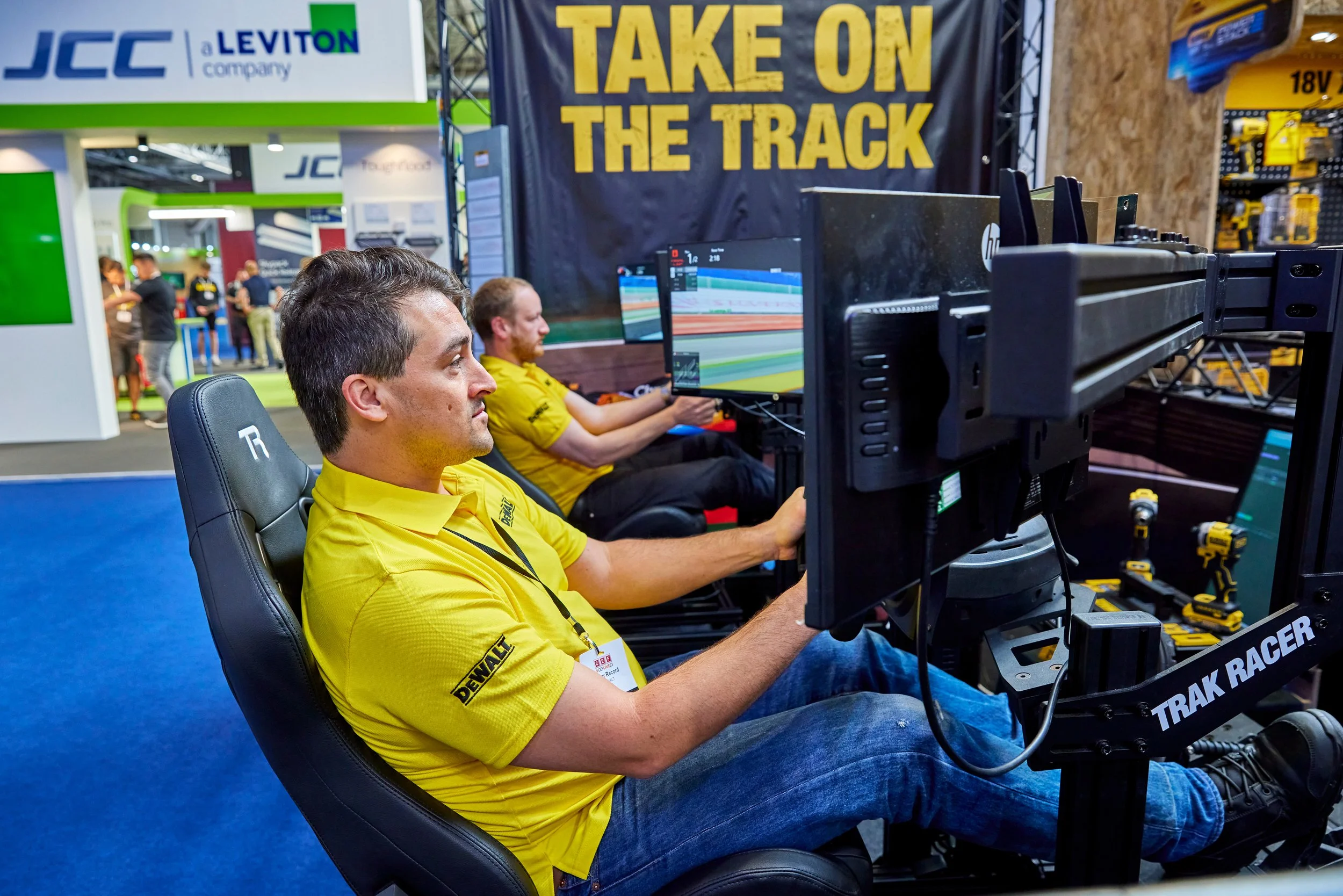 Two men in yellow shirts sitting in front of racing simulation monitors and equipment at a trade show. The background features banners and booths, including one with the slogan 'Take on the Track'.