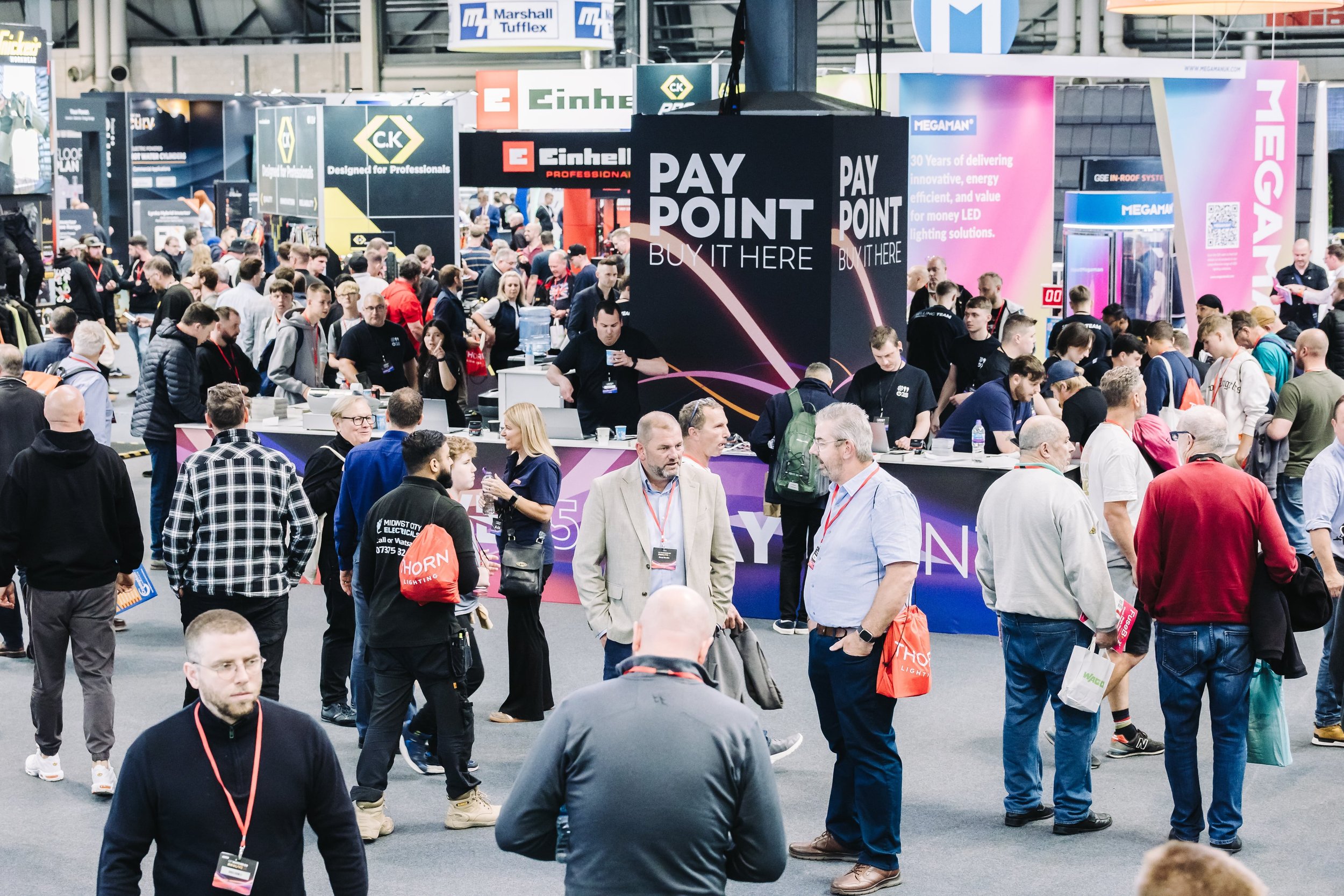 Trade show with people walking and interacting, booths with signs including "PAY POINT" and various brand banners in the background.
