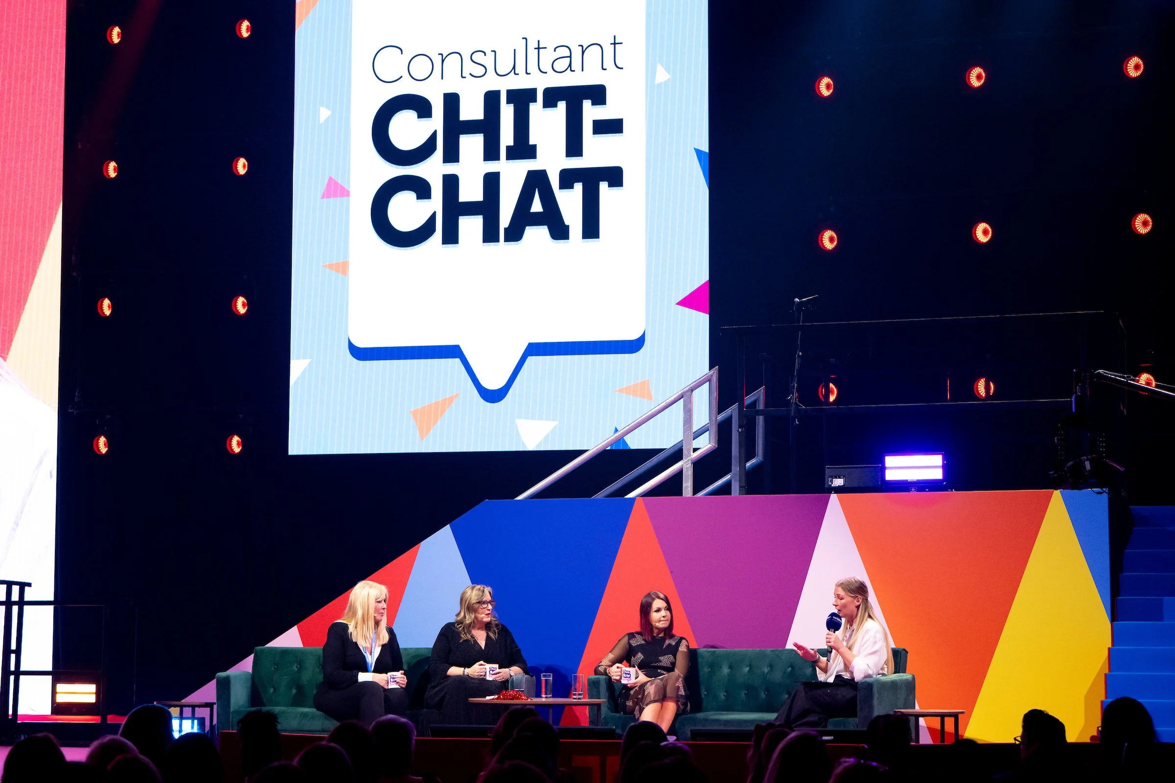 Women participating in a panel discussion on stage at a conference, with a large screen displaying 'Consultant Chit-Chat' behind them.