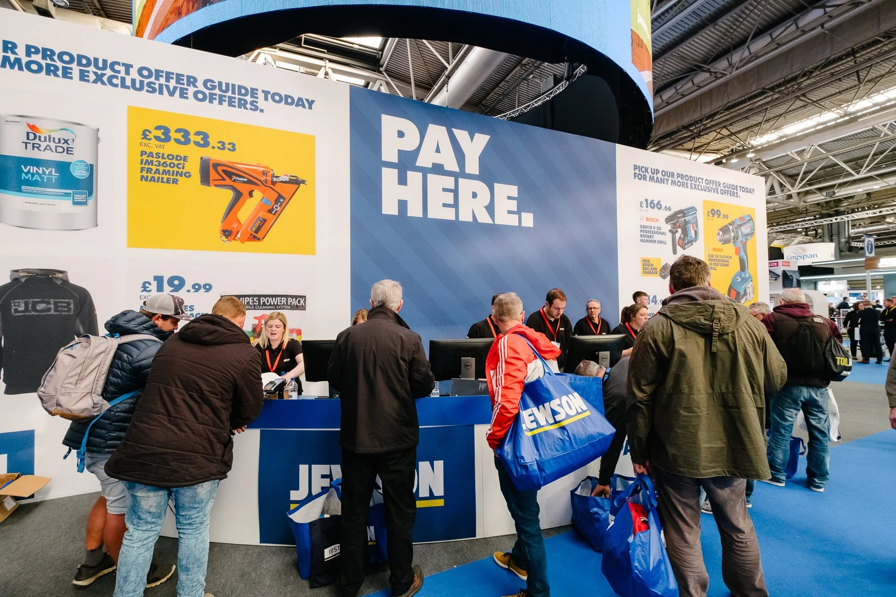 People at a booth with a large sign reading 'Pay Here' at a trade show. The booth has promotional posters for tools and paint, with customers waiting in line to check out. The setting is indoors with high ceilings and visible metal trusses.