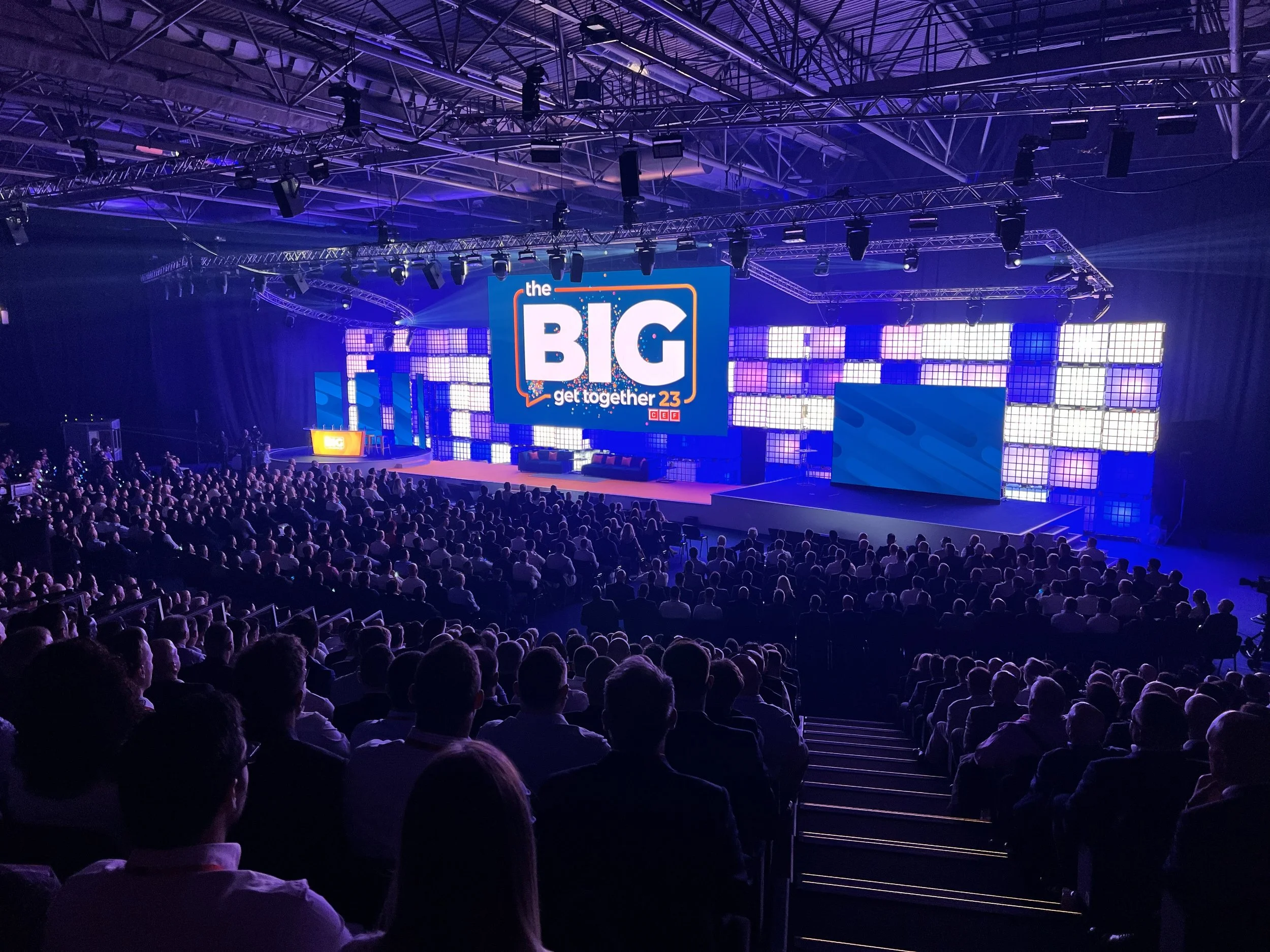 Large audience seated in a theater with a stage and a big screen displaying 'the BIG get together 23' with vibrant purple and blue lighting.