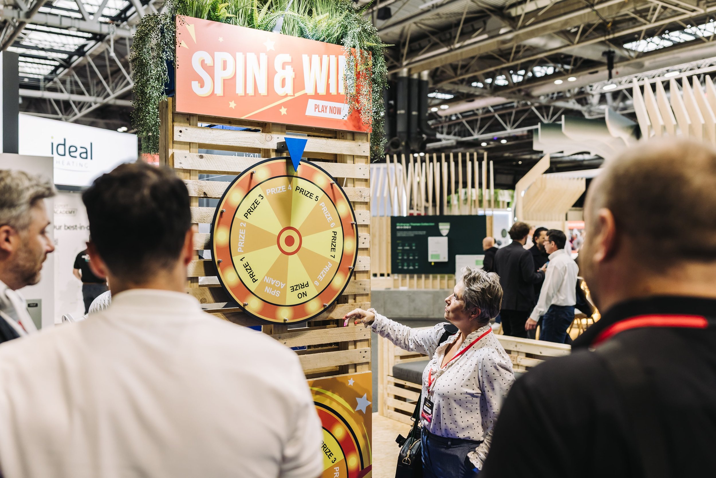 A woman with gray hair and glasses points at a spinning prize wheel at a booth called 'Spin & Win' with a sign that says 'Play Now.' Several people are gathered around, watching the wheel, in a busy indoor event or exhibition space with wooden struct