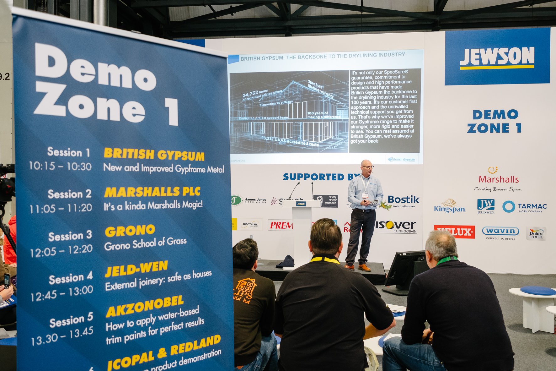 Presentation at a trade event on British Gypsum with a speaker standing on stage and a screen showing details about the company and its products. The audience is seated and watching.