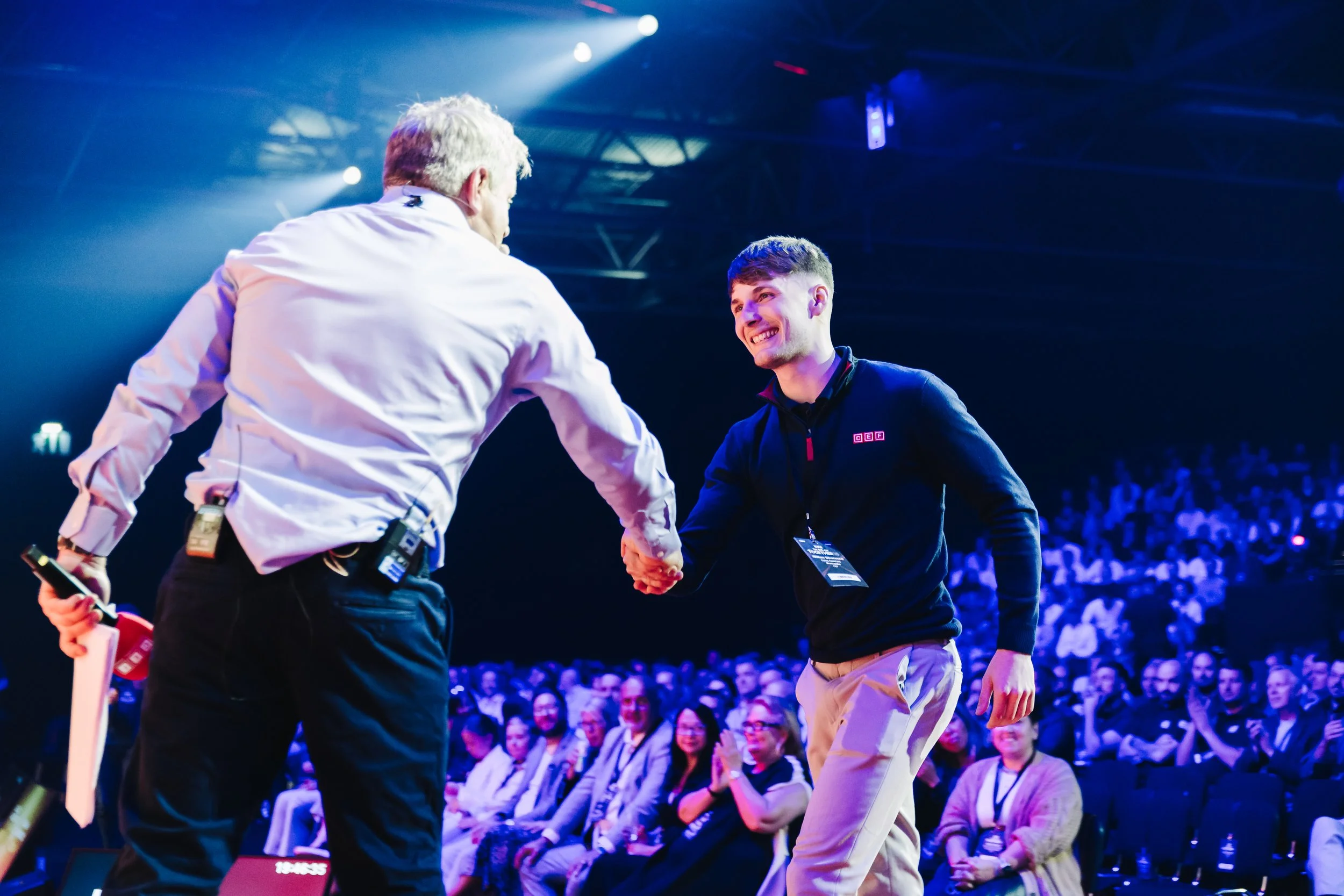 Two men shake hands on stage at a conference with an audience watching, one wearing a white shirt and the other a black jacket, in a large indoor venue with blue and purple lighting.
