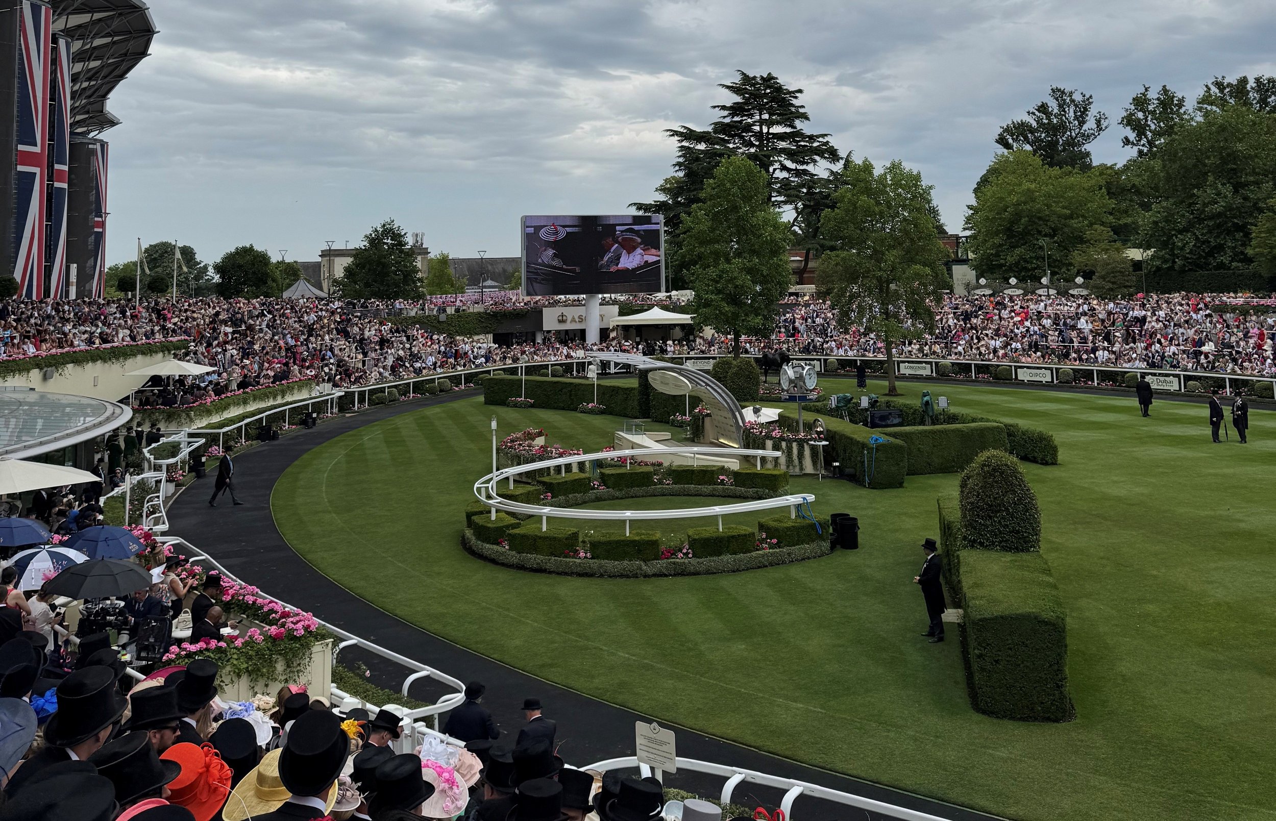 Crowd gathered at a horse racing track with lush green grass, flowers, and spectators in hats and umbrellas, overcast sky.
