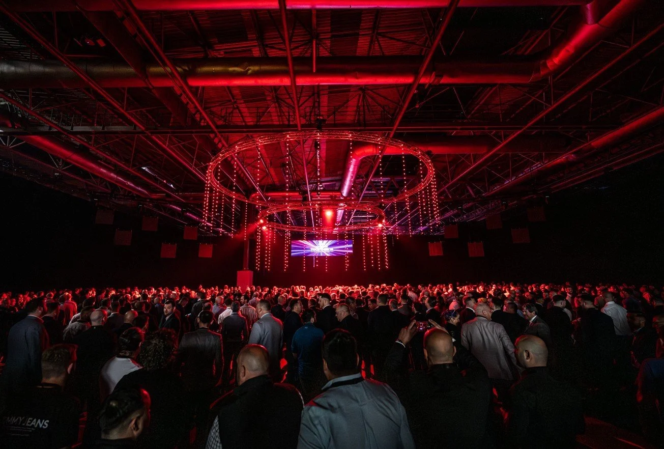 Large indoor concert or event venue filled with a crowd of people, illuminated by red stage lighting, with hanging LED lights and a central illuminated structure.