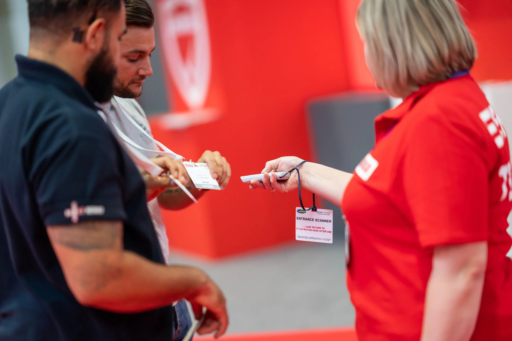 A woman in a red shirt scans tickets for two men, one with a beard and tattoos, holding their tickets while they wait to enter an event.