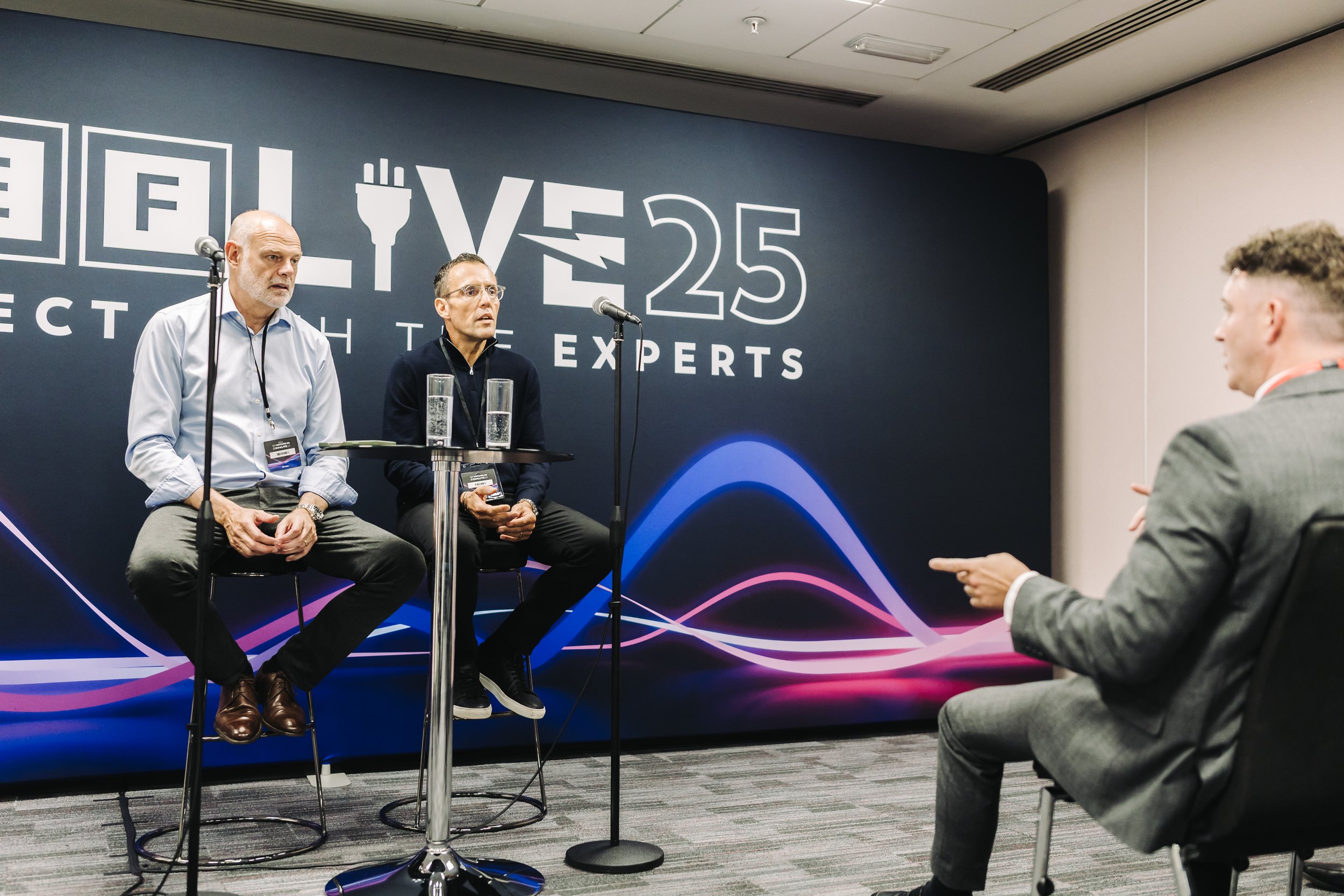 Two men sitting on high chairs with glasses of water in front of a dark blue wall with a logo that reads 'YES 25' and tagline 'Connect with the Experts'. They are in a panel discussion or interview setup, facing a man in a gray suit who is asking que
