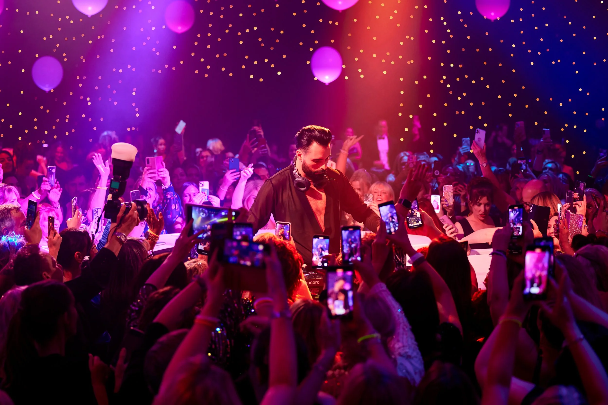 A DJ performing at a crowded music event with colorful lighting and people taking photos with their smartphones.