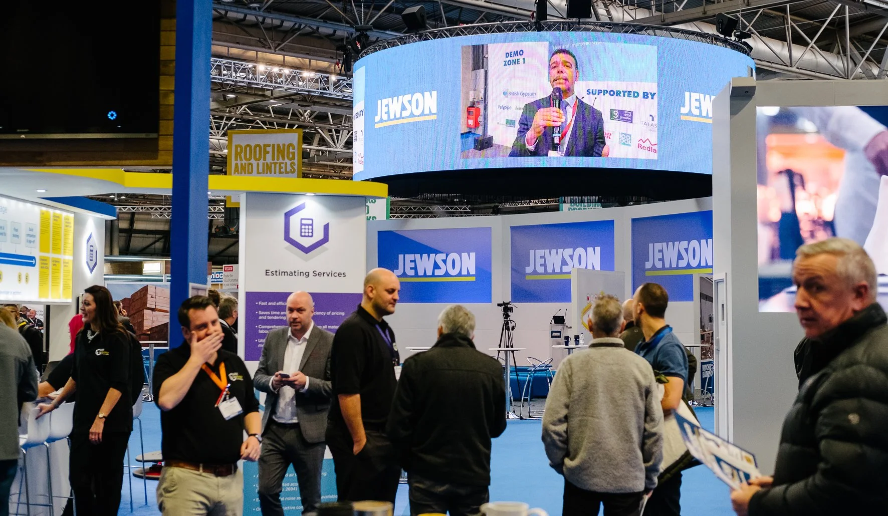 Industry trade show booth with large screens displaying a man speaking, the booth features signage for Jewson and Estimating Services, attendees are engaging and walking around the exhibition area.