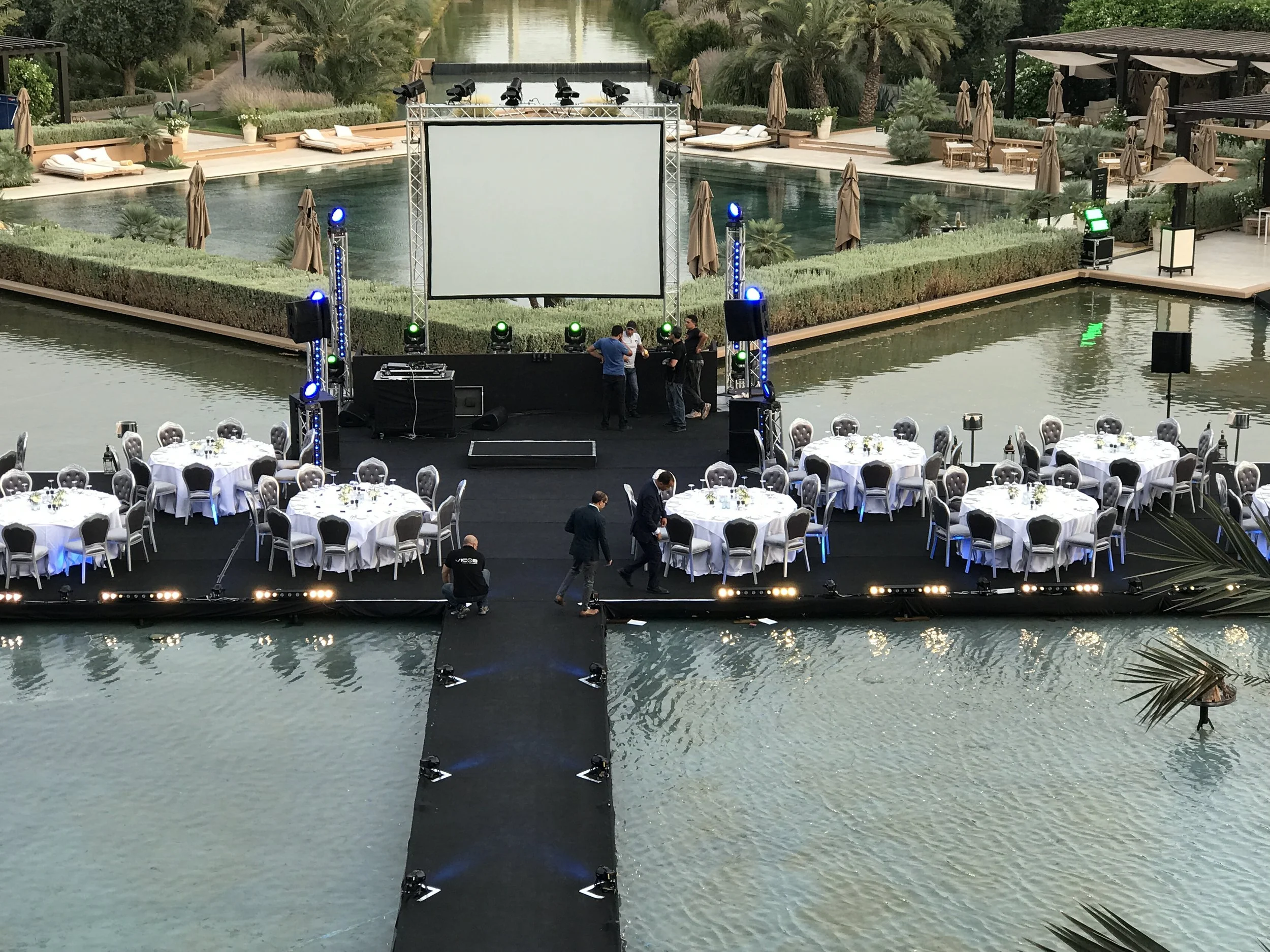 An outdoor event setup with round tables covered with white tablecloths and black chairs on a floating platform over water. There is a stage with lighting, sound equipment, and a large blank screen, with people preparing for an event in the early evening.