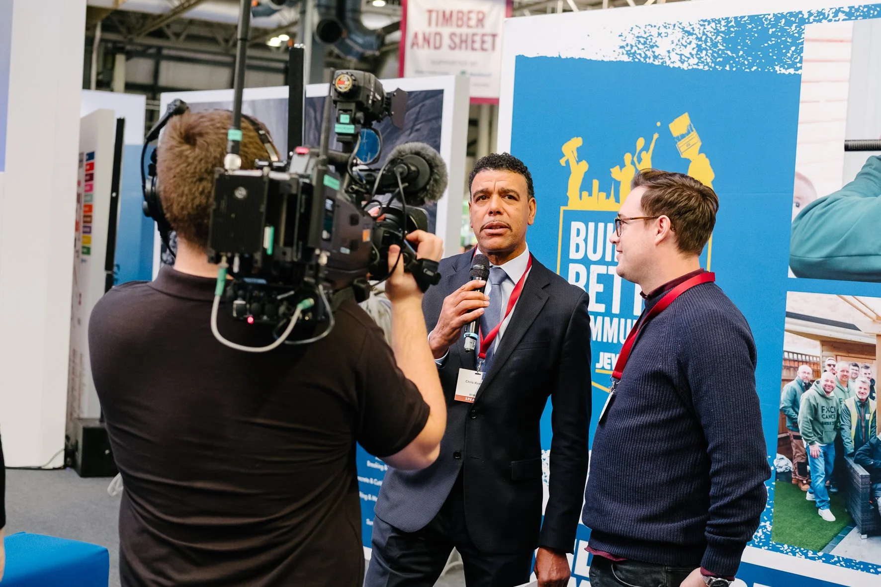 A man in a suit and tie holding a microphone being interviewed by a cameraman at an indoor event, with a blue banner in the background featuring the words 'Build Better Communities' and images of people raising their hands.