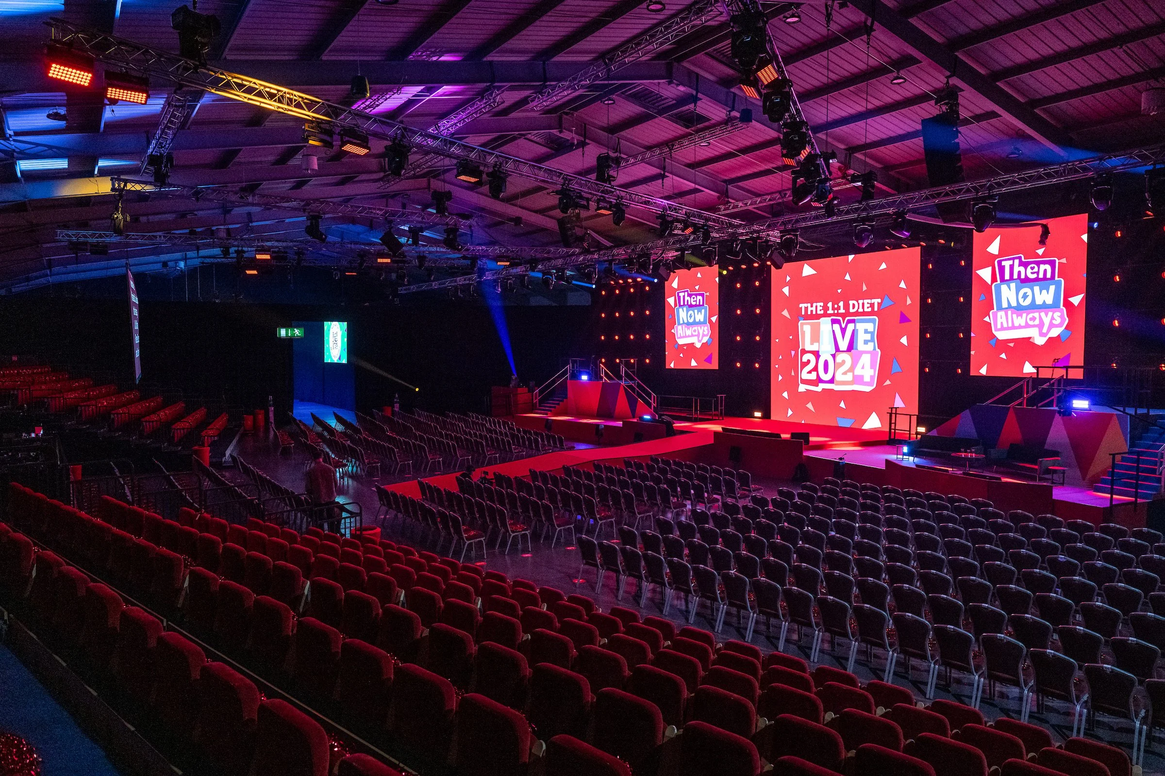 Empty conference or concert hall with a stage, large screens displaying colorful graphics that say 'The 1:1 Diet Live 2024,' and comfortable red and black chairs arranged in rows facing the stage. The hall has a modern setup with lighting and rigging.