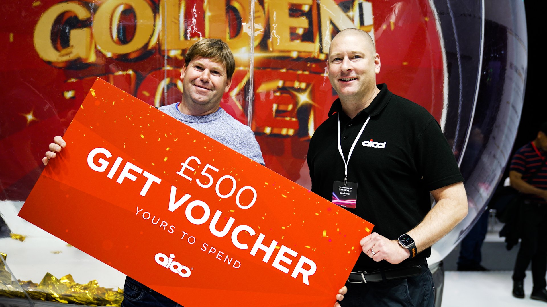 Two men smiling, holding a large red gift voucher with white text that reads £500 GIFT VOUCHER. They are standing in front of a large background display that says 'GOLDEN'.