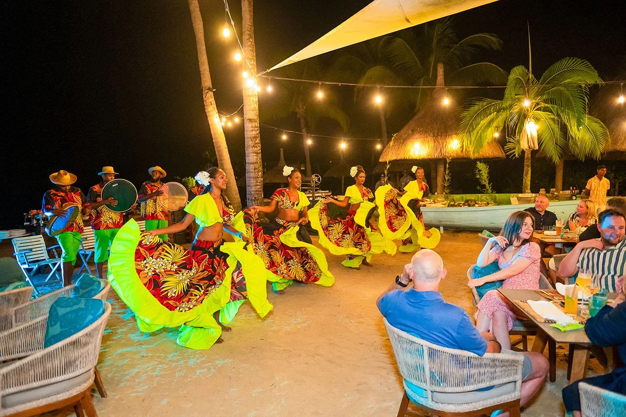 Hula dancers performing in front of an outdoor dining area at night, illuminated by string lights, with tropical trees and a thatched-roof hut in the background.