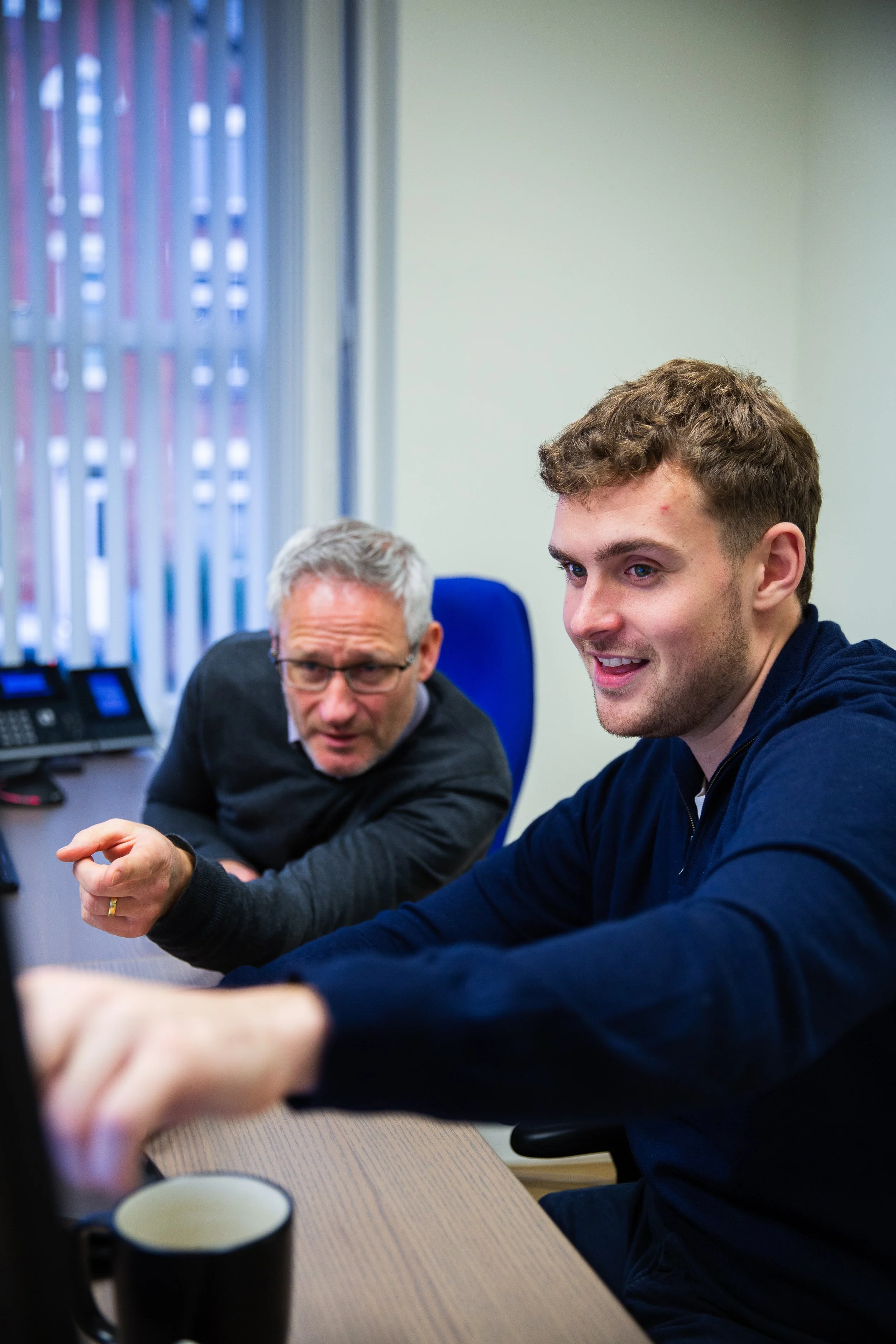 Two men in an office engaged in a discussion, with one pointing at a screen and the other reacting attentively.