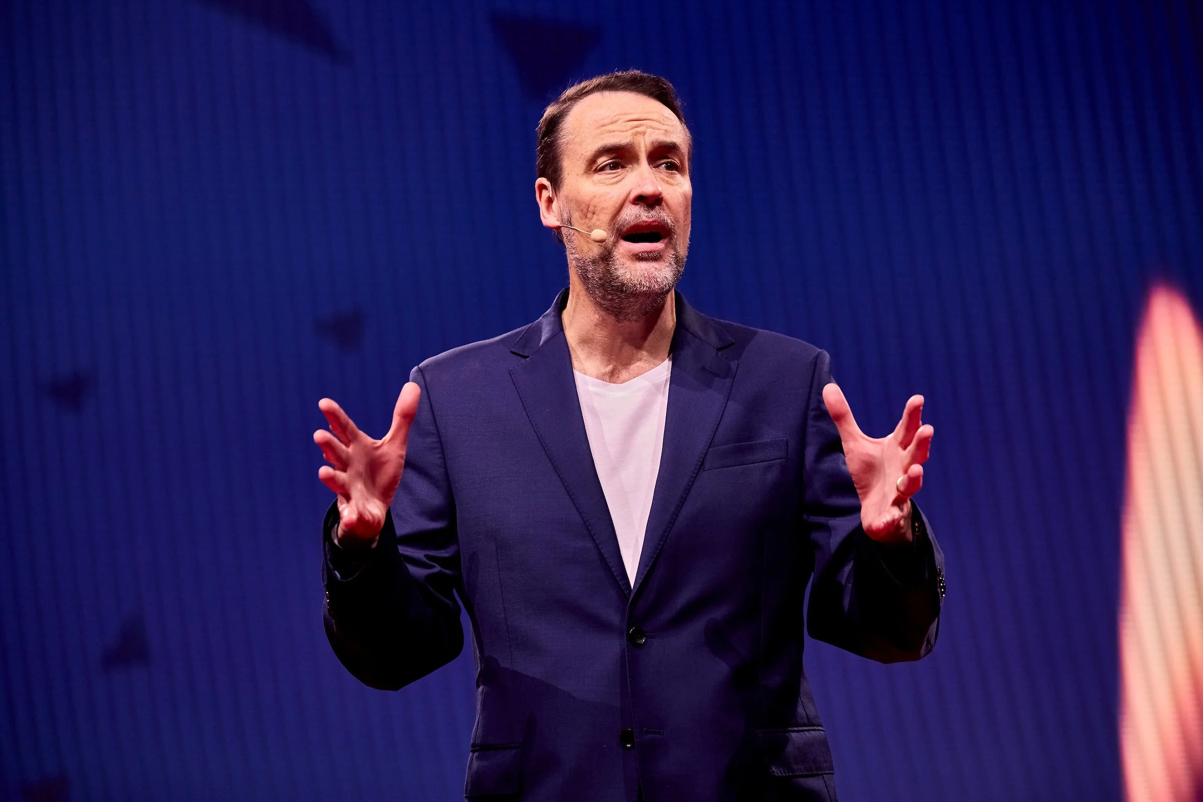 A man with dark hair and a beard wearing a navy blazer and white shirt speaking passionately on stage with a blue background.