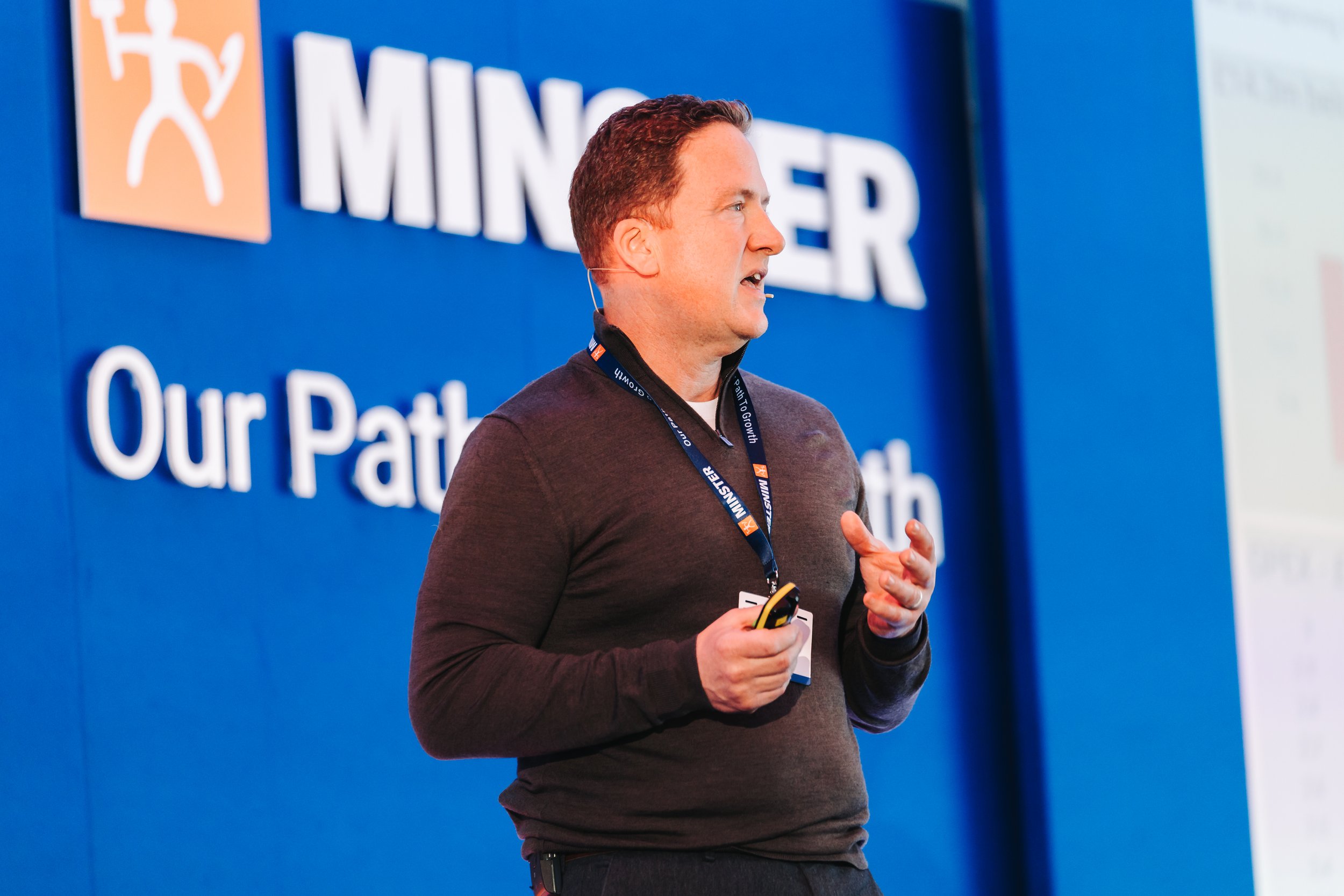 A man giving a presentation, standing in front of a blue backdrop with the text 'MINES' and 'Our Pat' visible, holding a remote control.