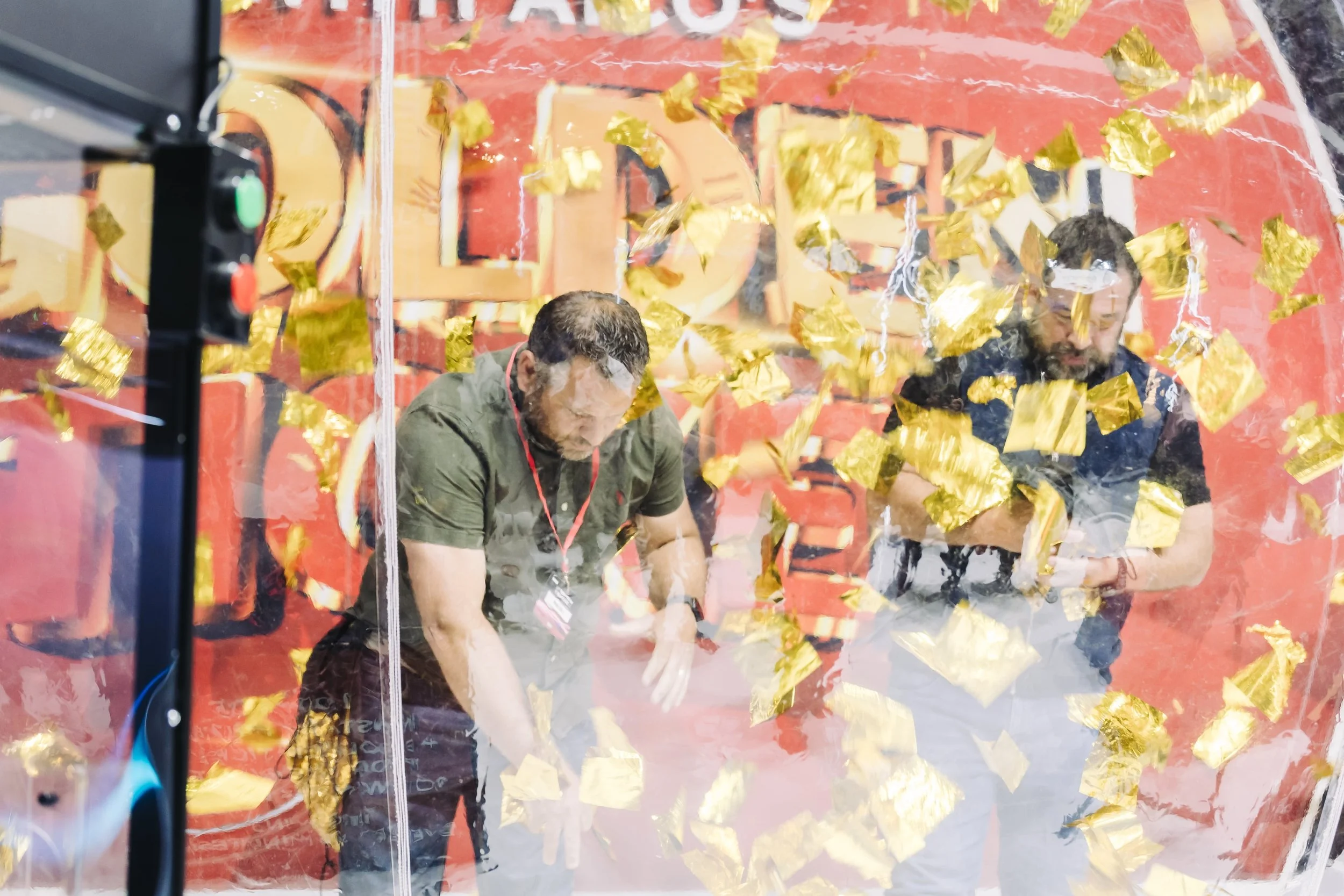 Two men leaning during an indoor event, seen through a clear cover filled with floating gold confetti.