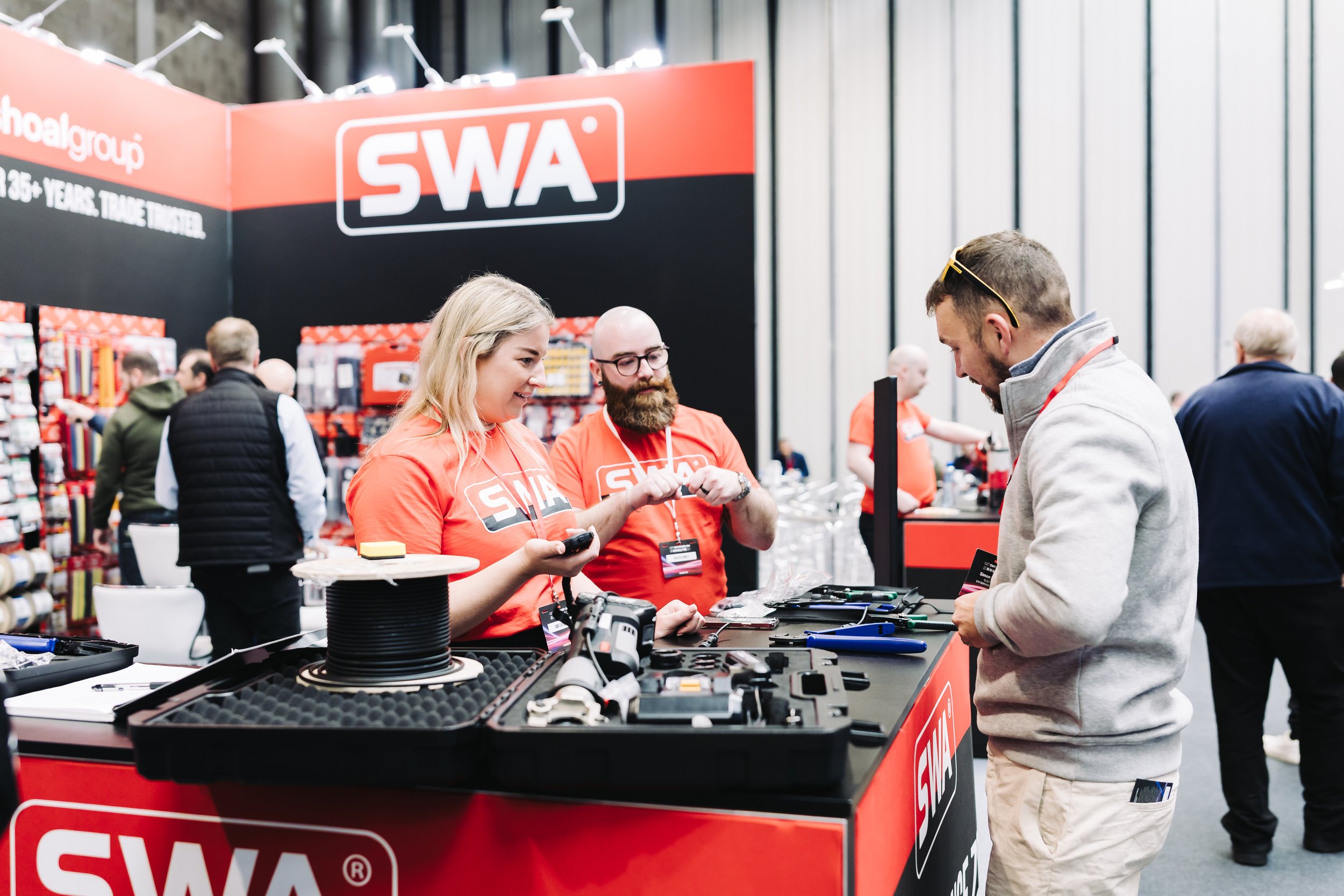 At a trade show booth for SWA, two representatives in red shirts are assisting a man in a gray sweatshirt, with tools and equipment laid out on the table.