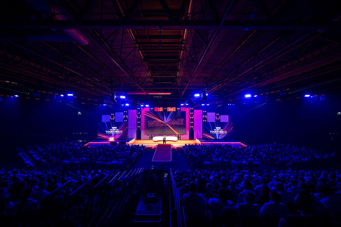 Large indoor conference or event with a stage, audience, and colorful lighting, displaying the word 'WELCOME' on the main screen.