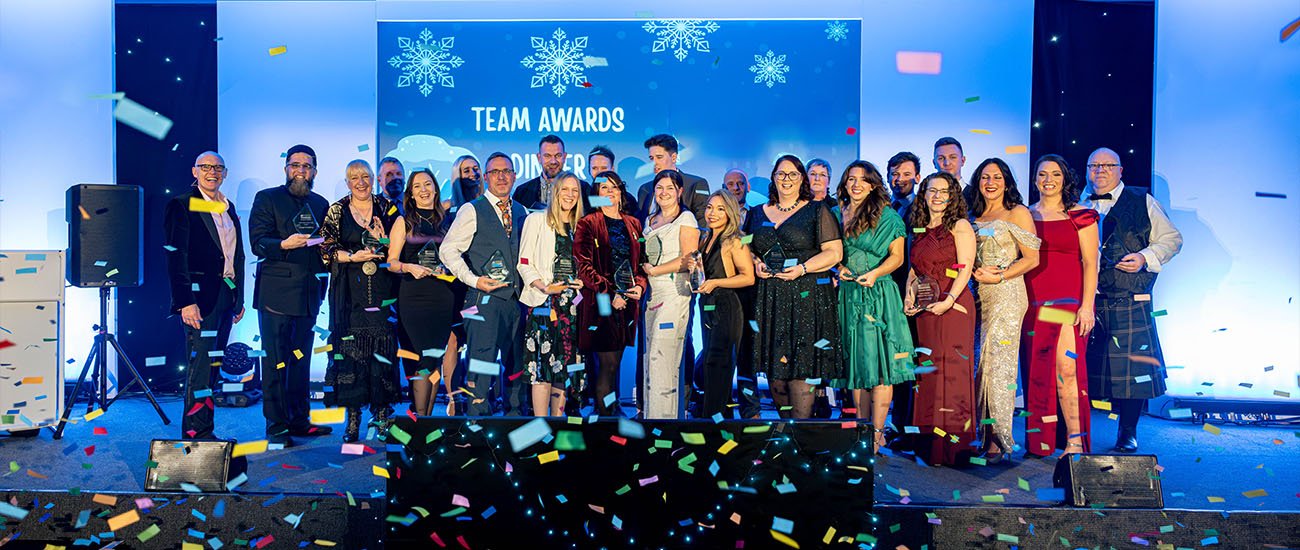 Group of people dressed in formal attire on stage holding awards, with confetti falling, at an event with a screen displaying 'Team Awards Ceremony' and snowflake decorations.