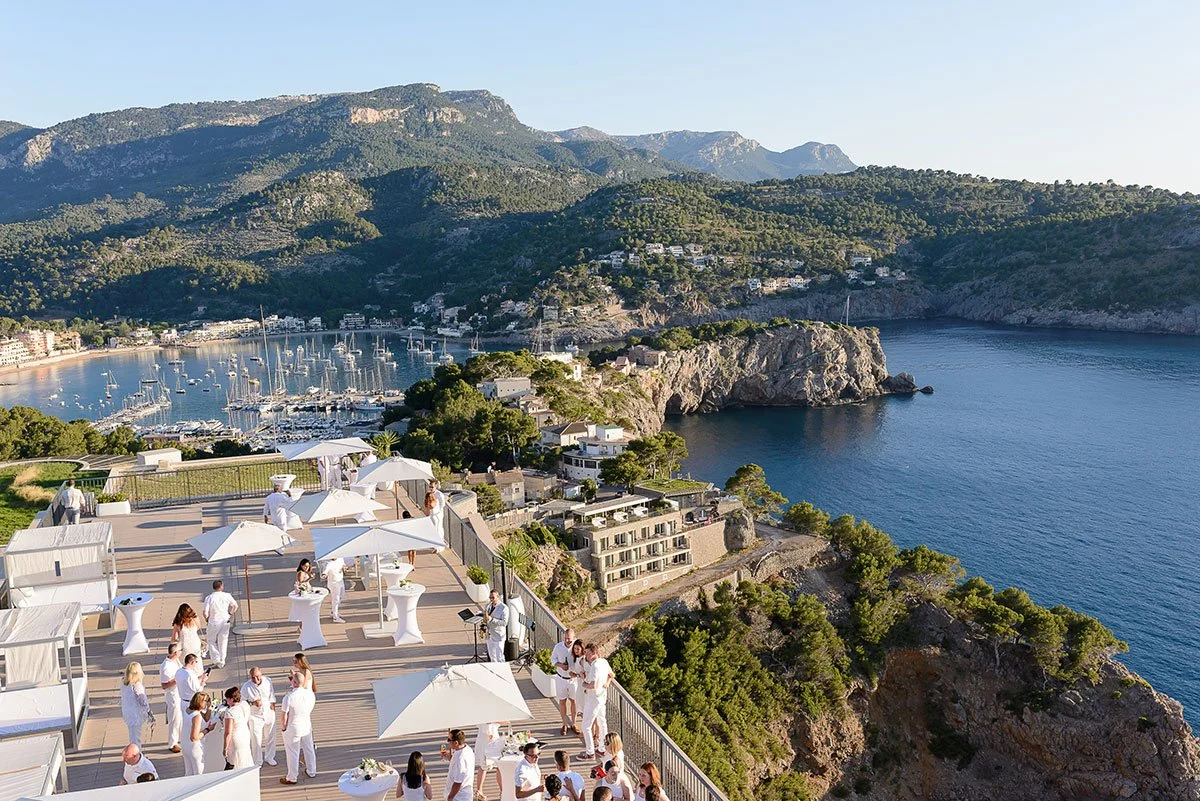 People dressed in white attending an outdoor event on a terrace overlooking a bay with sailboats, surrounded by green hills and mountains.