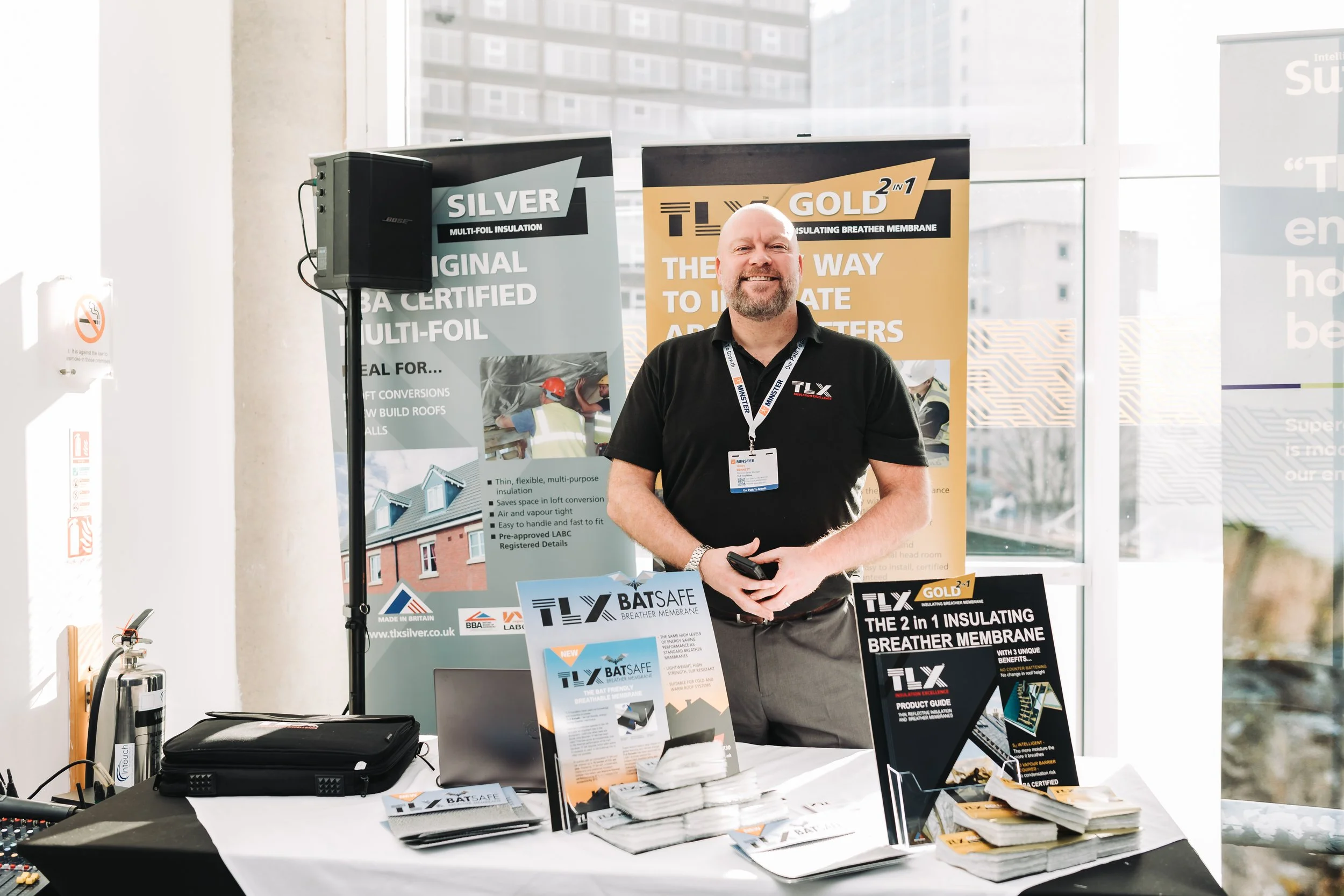 A man standing behind a booth with promotional materials for insulation products, including posters and brochures, inside a building with large windows.