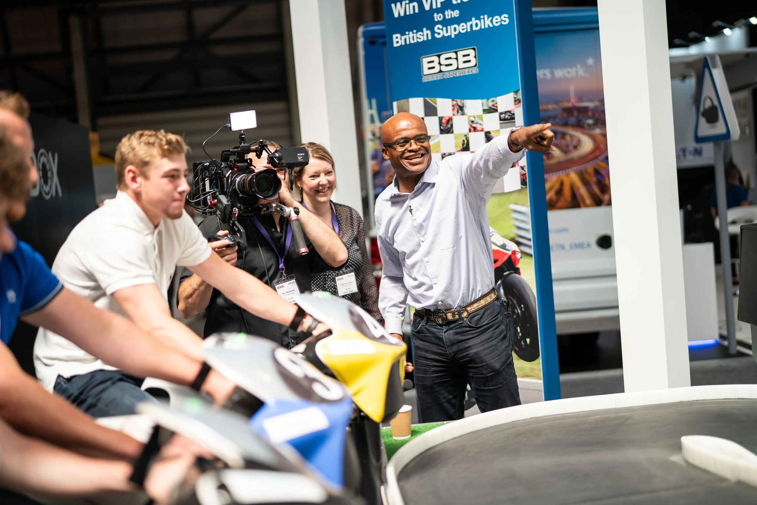 A man in a white shirt pointing and smiling while speaking at a racing event, surrounded by a group including young men on slot cars, a camera operator, and a woman, at an indoor exhibition with a blue banner that reads 'Win VIP tickets to the Britis