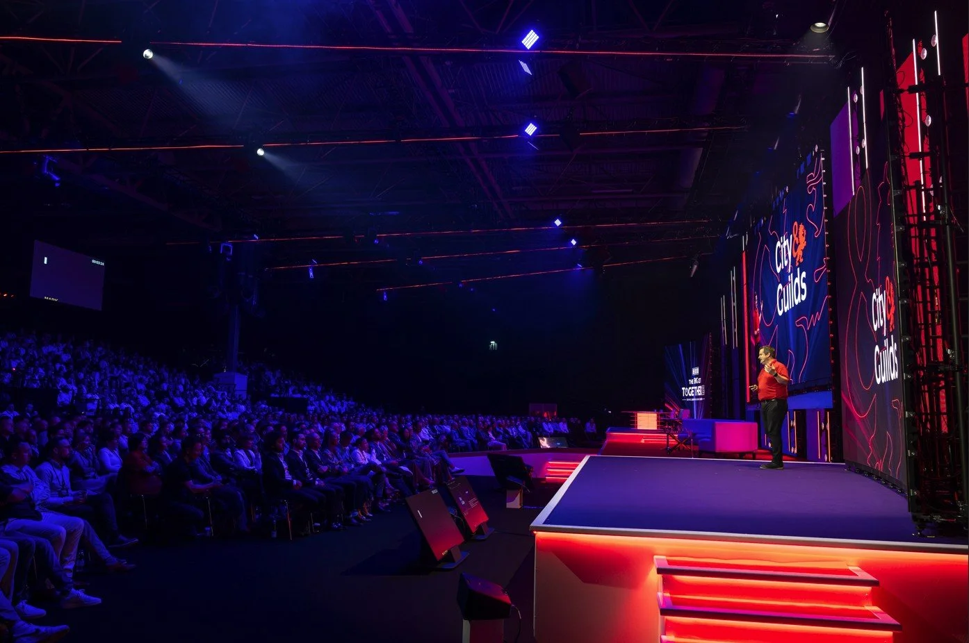 A speaker on stage delivering a presentation at a conference called City Guides, with a large audience seated in front, illuminated by colorful stage lighting.
