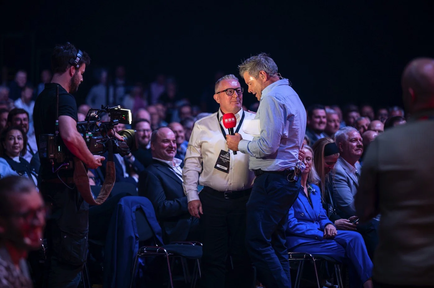 A man interviewing another man holding a red microphone in front of an audience at a conference.