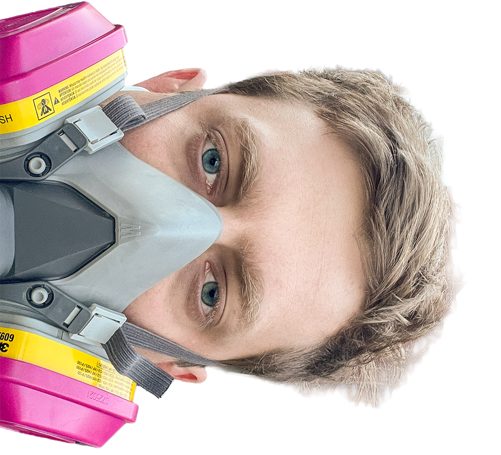 A man wearing a gray respirator mask with pink filters, looking at the camera with blue eyes and light brown hair.