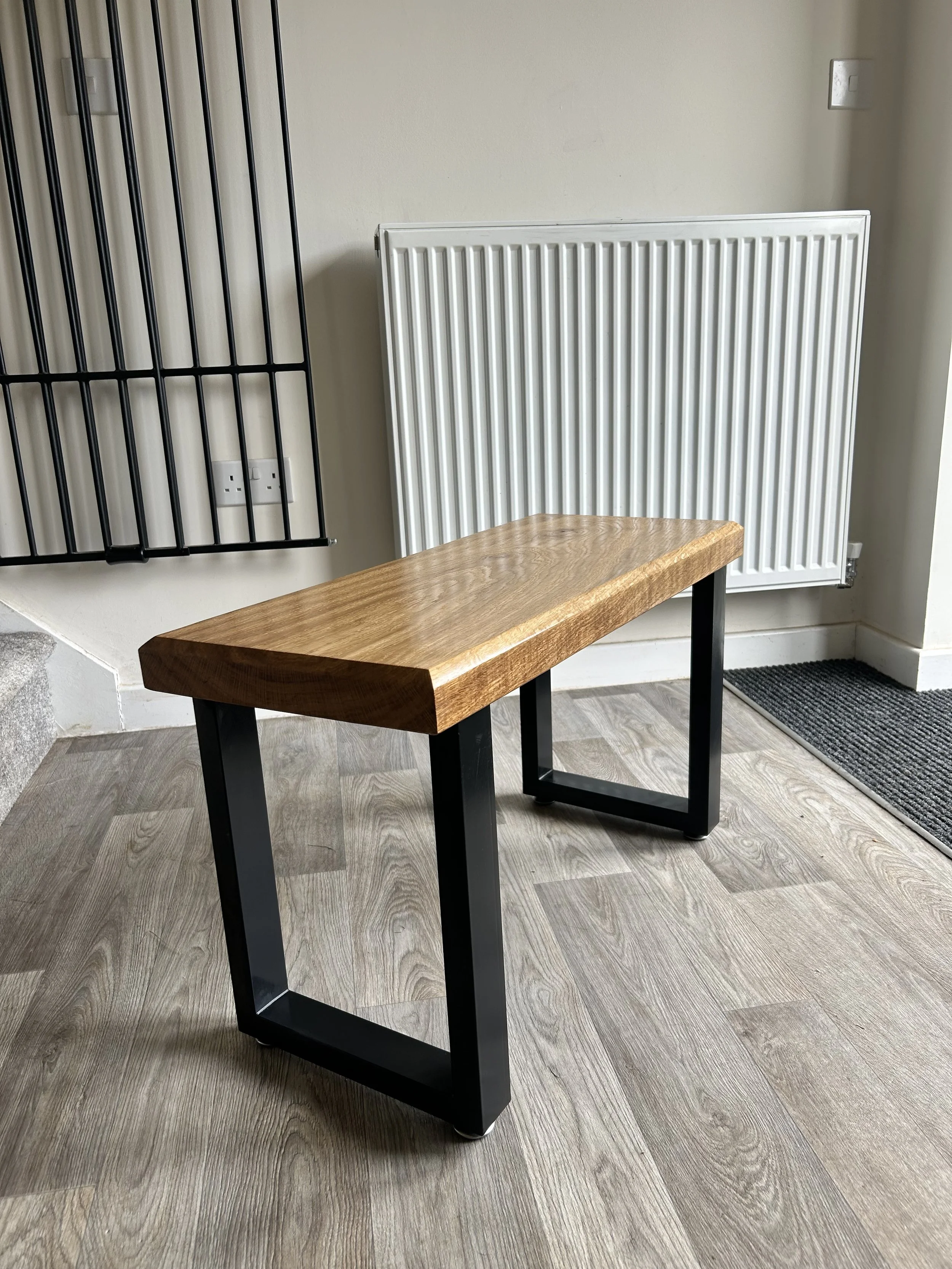 Wooden bench with black metal legs in a room with wooden flooring, a white radiator, a black wall-mounted shelf, and a gray doormat.