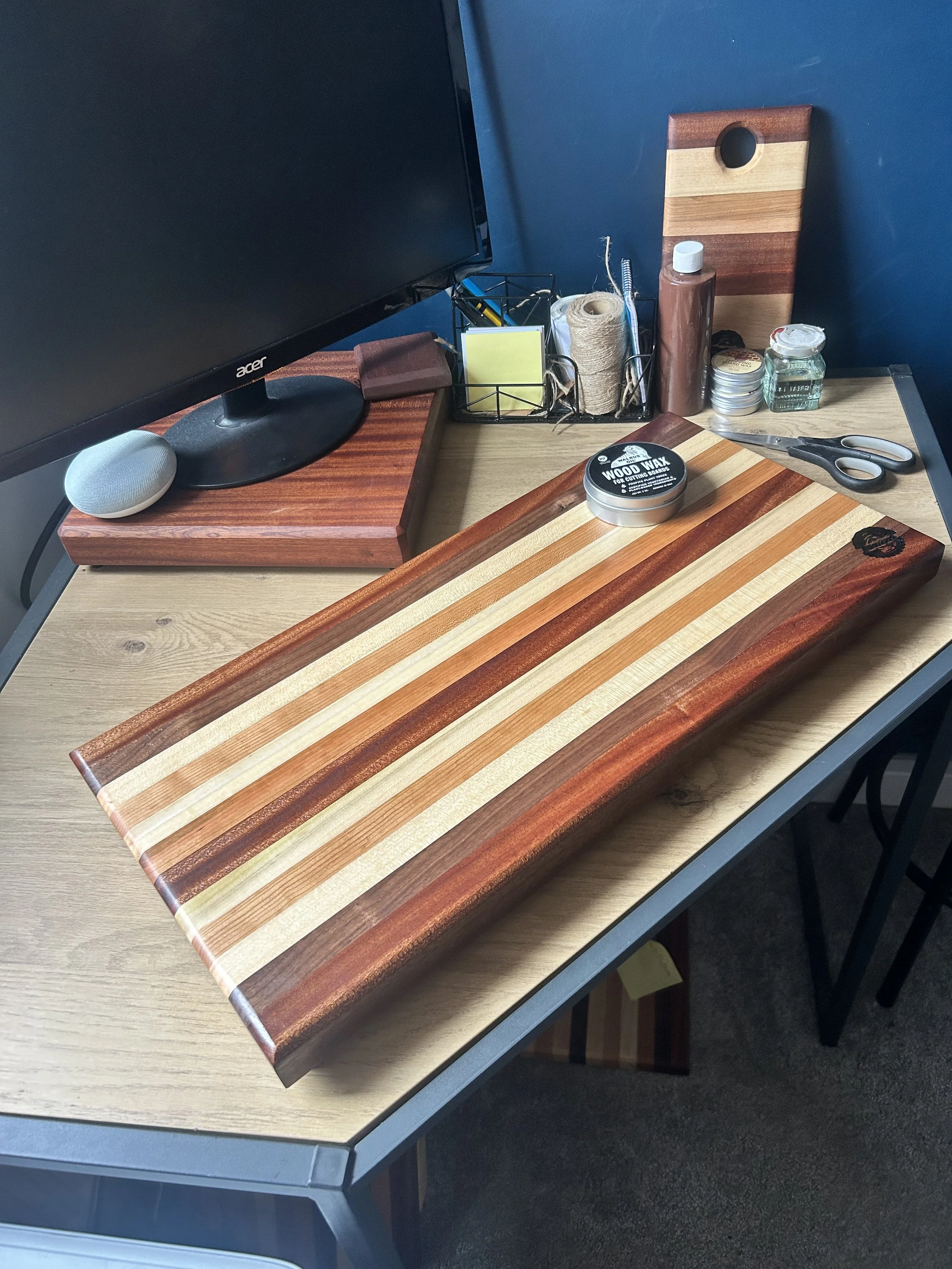 A wooden cutting board with alternating light and dark stripes on a desk, with scissors, a container of woodwax, and other small items in the background.