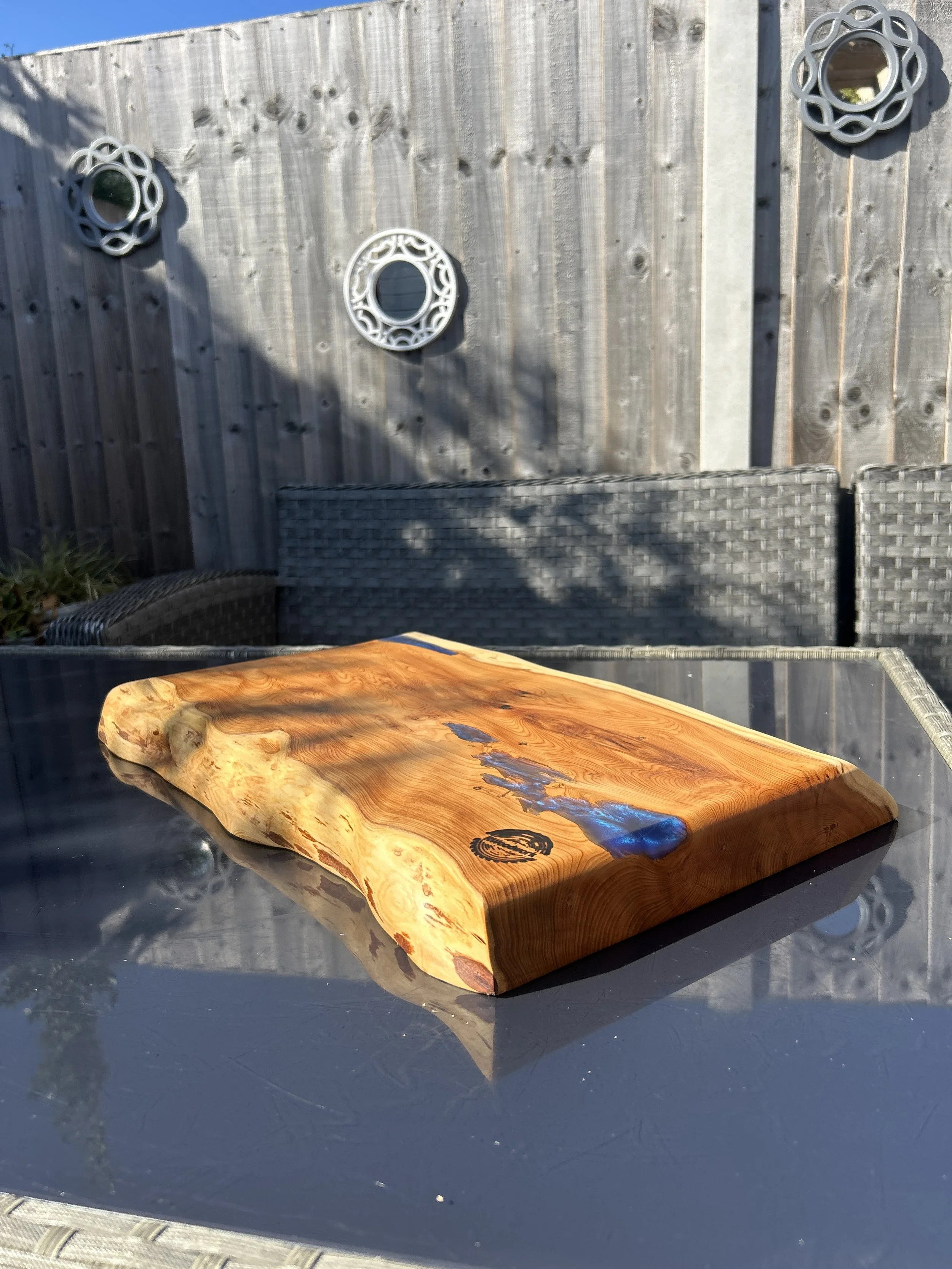 Wooden cutting board with a resin inlay on a glass outdoor table, with a gray wicker patio set and a wooden fence with decorative mirrors in the background.