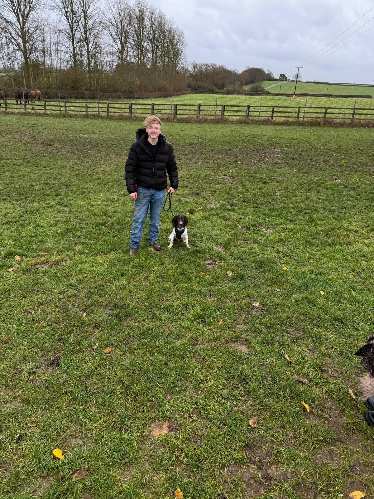 A young man in a black puffer jacket and blue jeans holding a leash attached to a small black and white dog, standing on a grassy field with a wooden fence and trees in the background.
