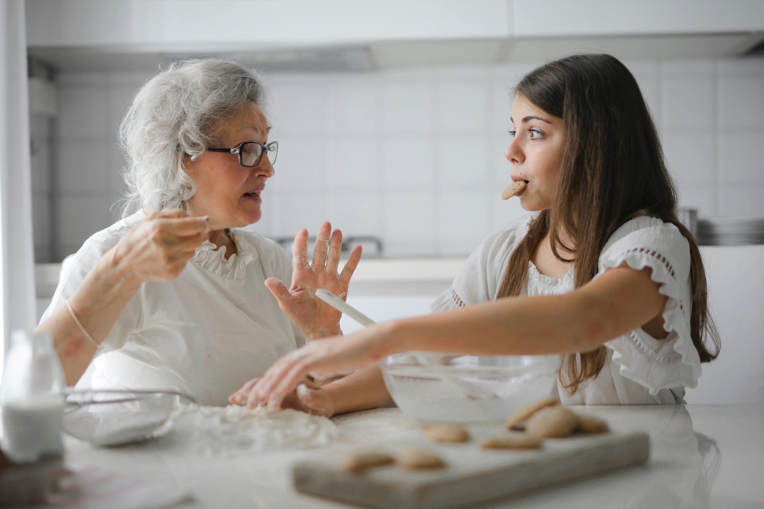 An elderly woman with gray hair and glasses talking to a young girl with long brown hair, who is holding a cookie in her mouth. They are in a kitchen with a white countertop, and there are cookies on a baking tray in front of them.