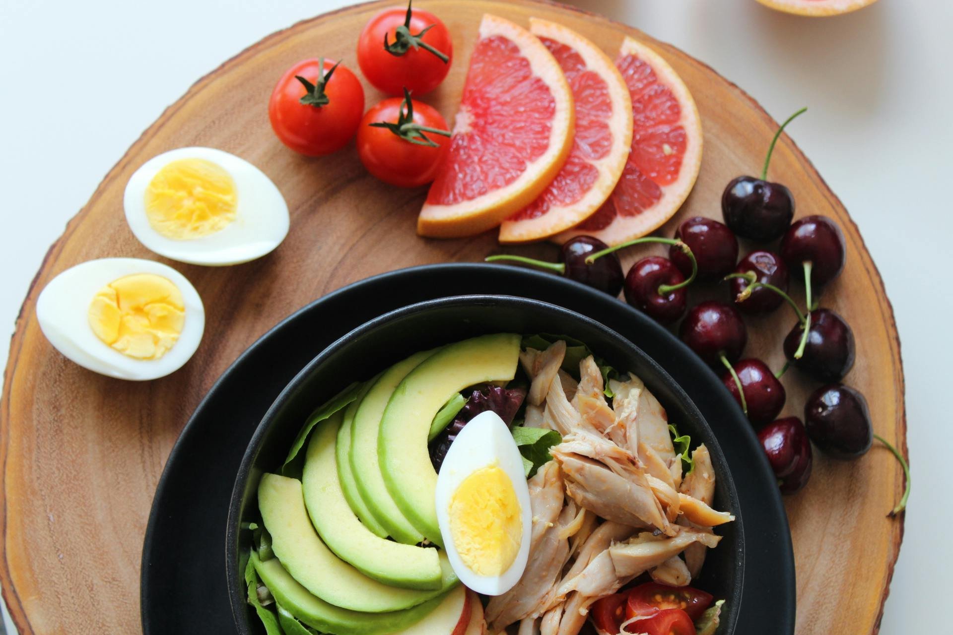 A wooden platter with cherry tomatoes, grapefruit slices, cherries, and a bowl of salad containing avocado, boiled eggs, shredded chicken, cherry tomatoes, and greens.