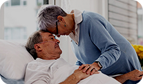 An elderly man in a hospital bed with a woman leaning over him, holding his hand, in a caring moment.