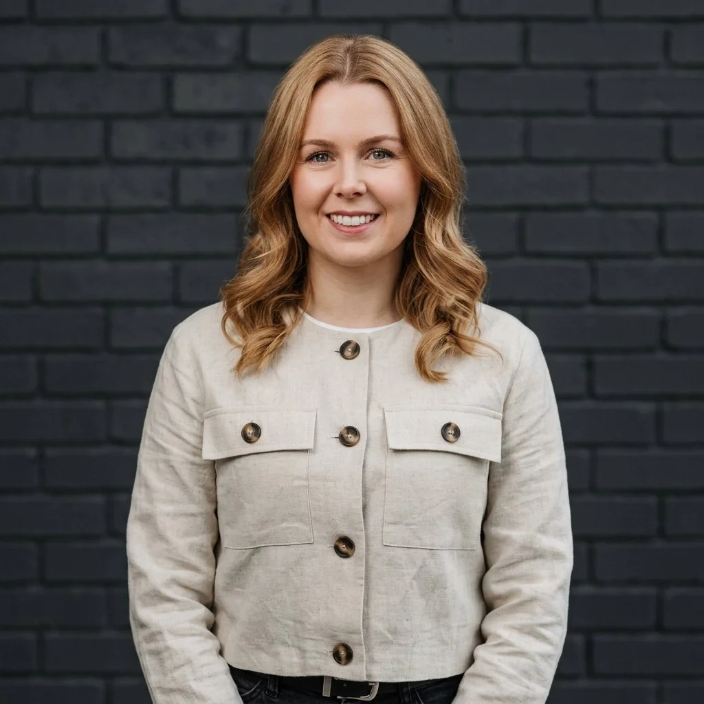 Smiling woman with wavy blonde hair wearing a light-colored jacket with large buttons, standing against a dark brick wall.