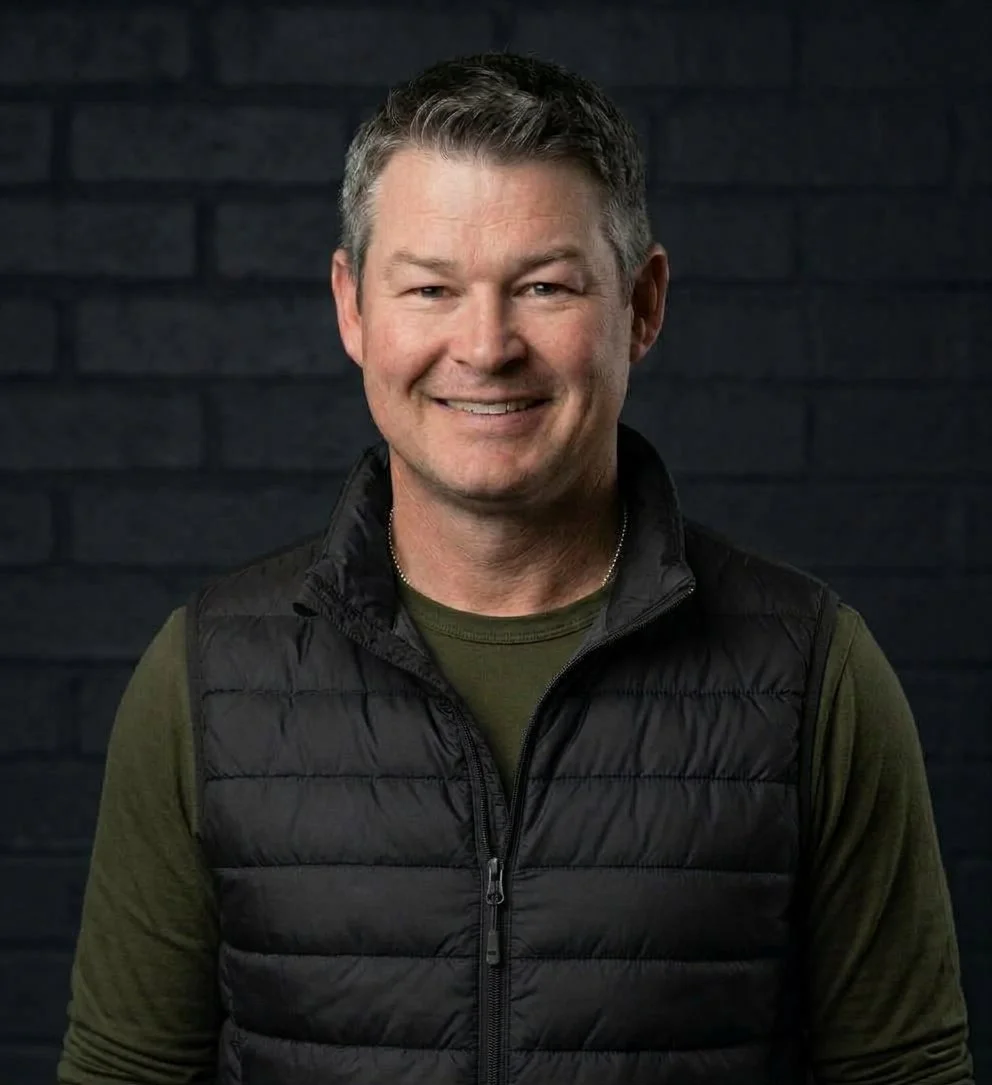 Smiling man with short dark hair and gray highlights, wearing a black quilted vest over a green shirt, in front of a dark brick wall.