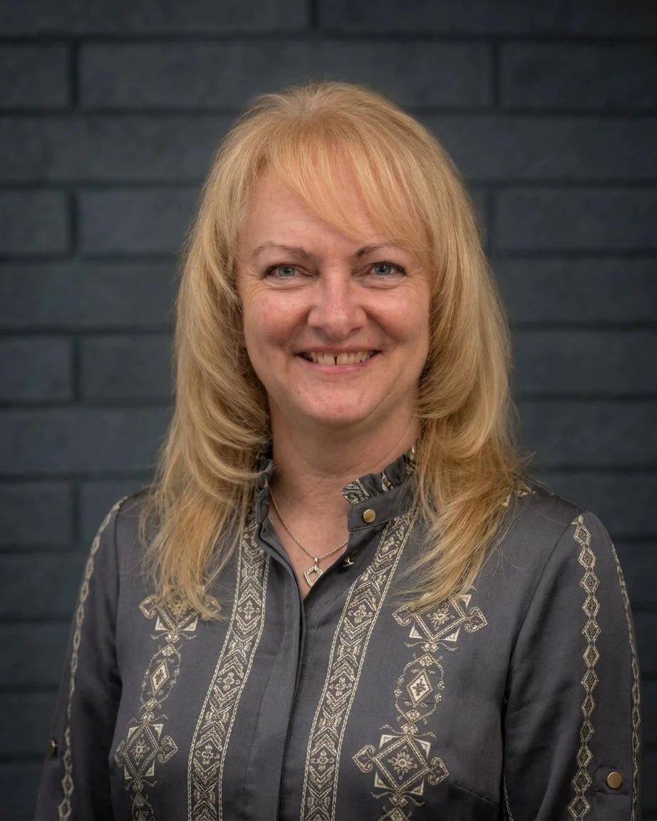 Portrait of a smiling woman with blonde hair, wearing a gray embroidered blouse, standing against a gray brick wall.