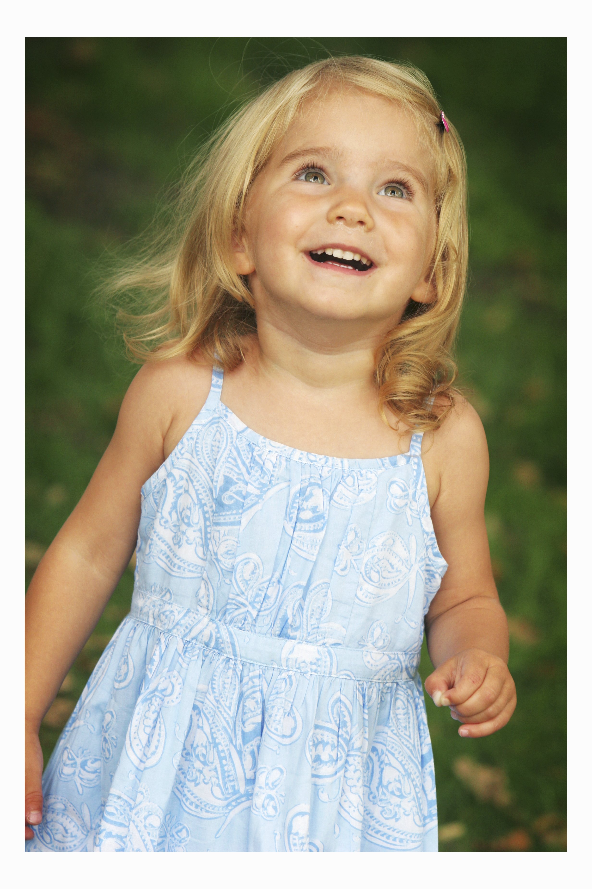 A young girl with blonde hair, wearing a light blue patterned dress, smiling and looking up outdoors with a blurred green background.