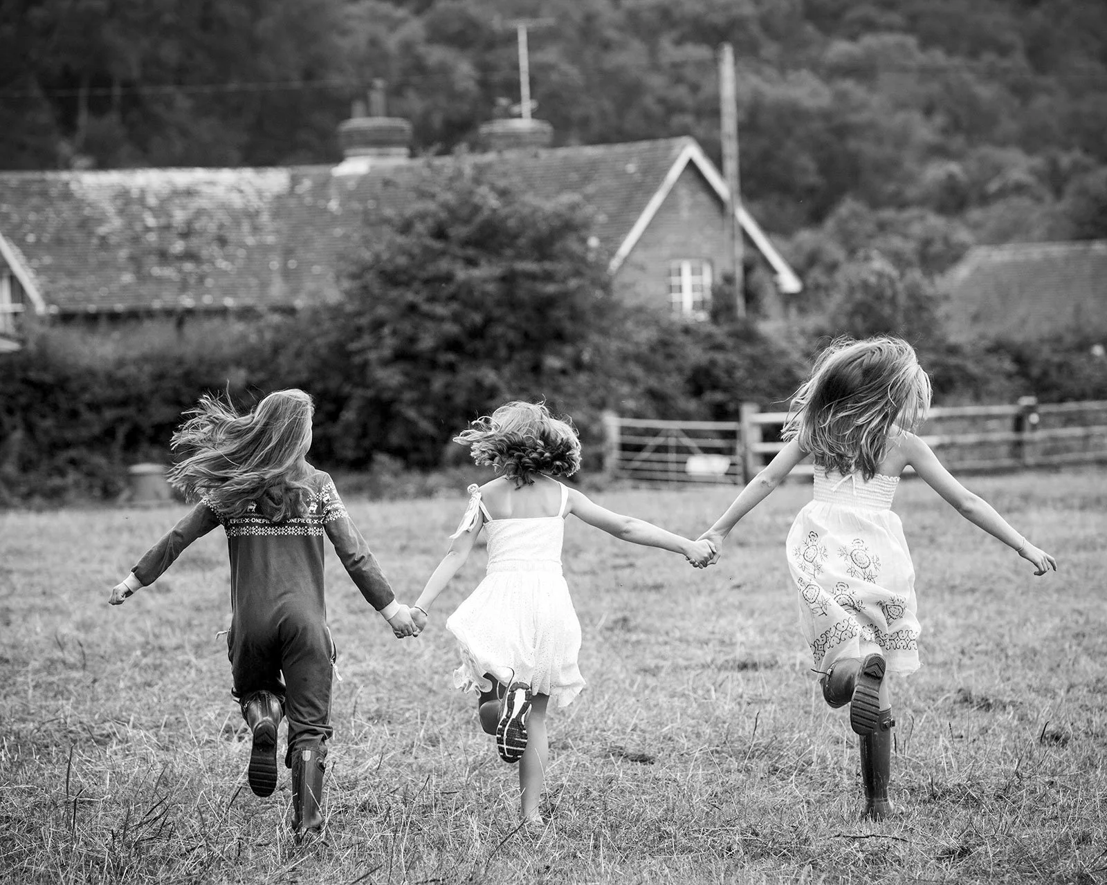 Three young girls holding hands and running in a grassy field, seen from behind, in black and white.