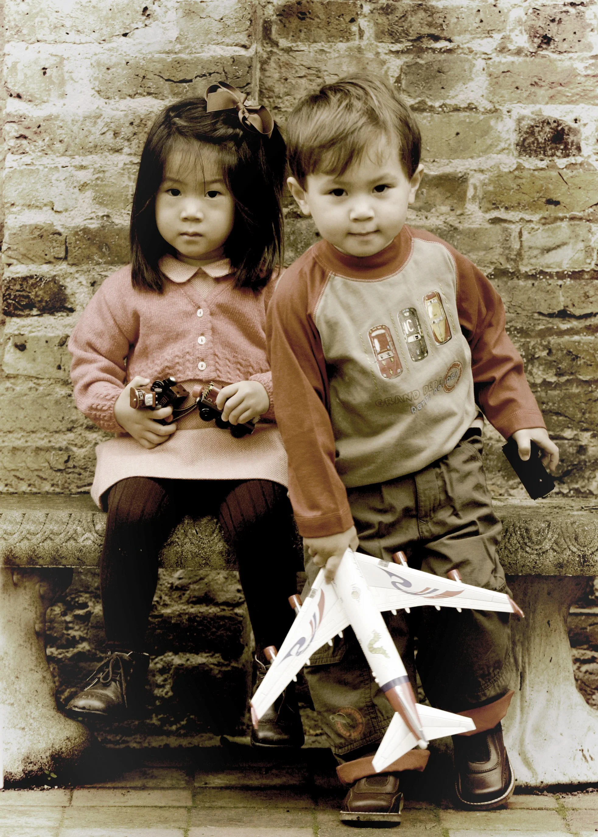 Two young children, a girl and a boy, sit on a stone bench against a brick wall. The girl, with long dark hair and a large bow, holds binoculars. The boy, with short light hair, holds a toy airplane and a small black object.